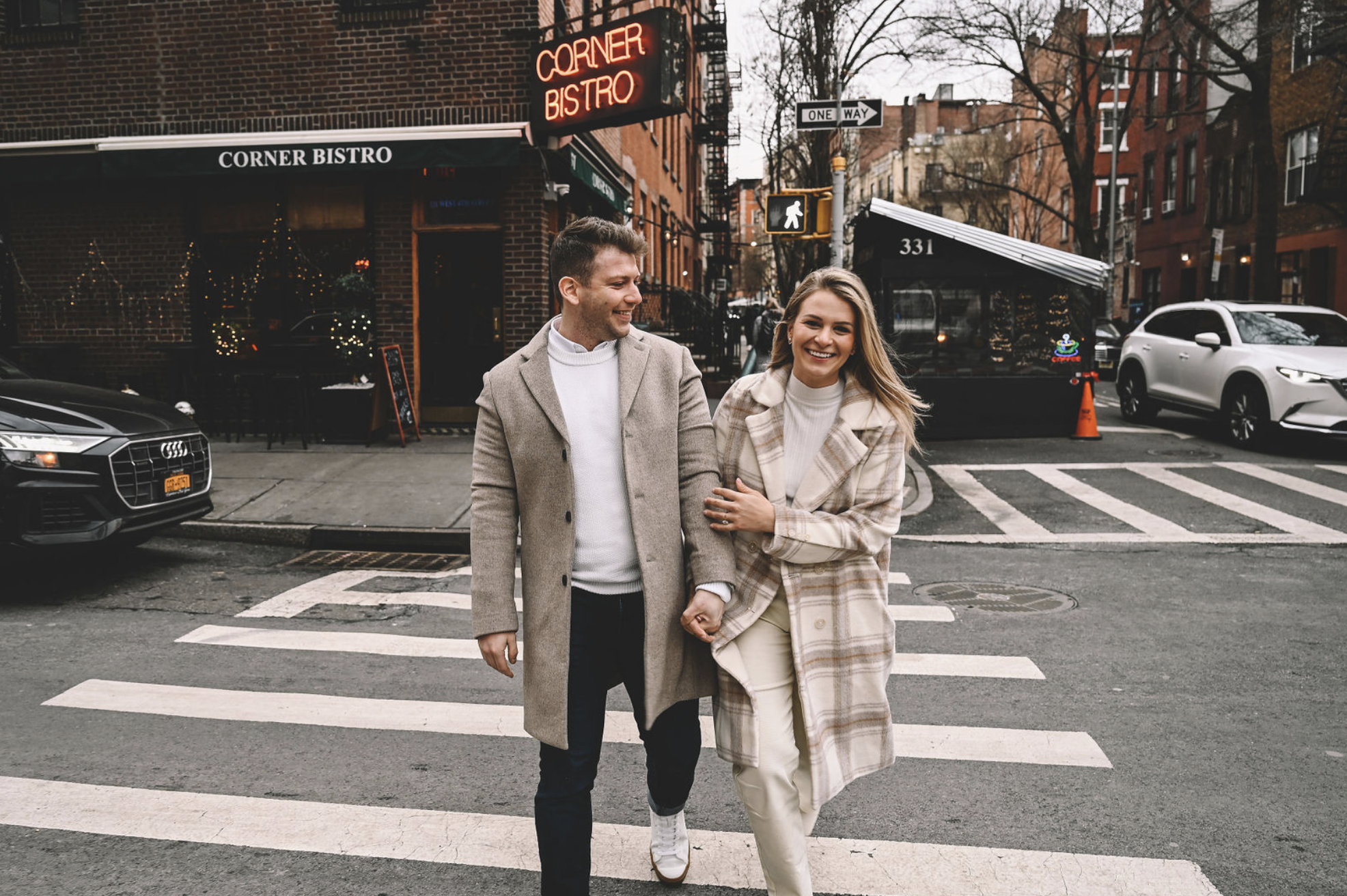 A smiling man and woman walking arm in arm across a city street at dusk, with a bistro and various cars in the background.