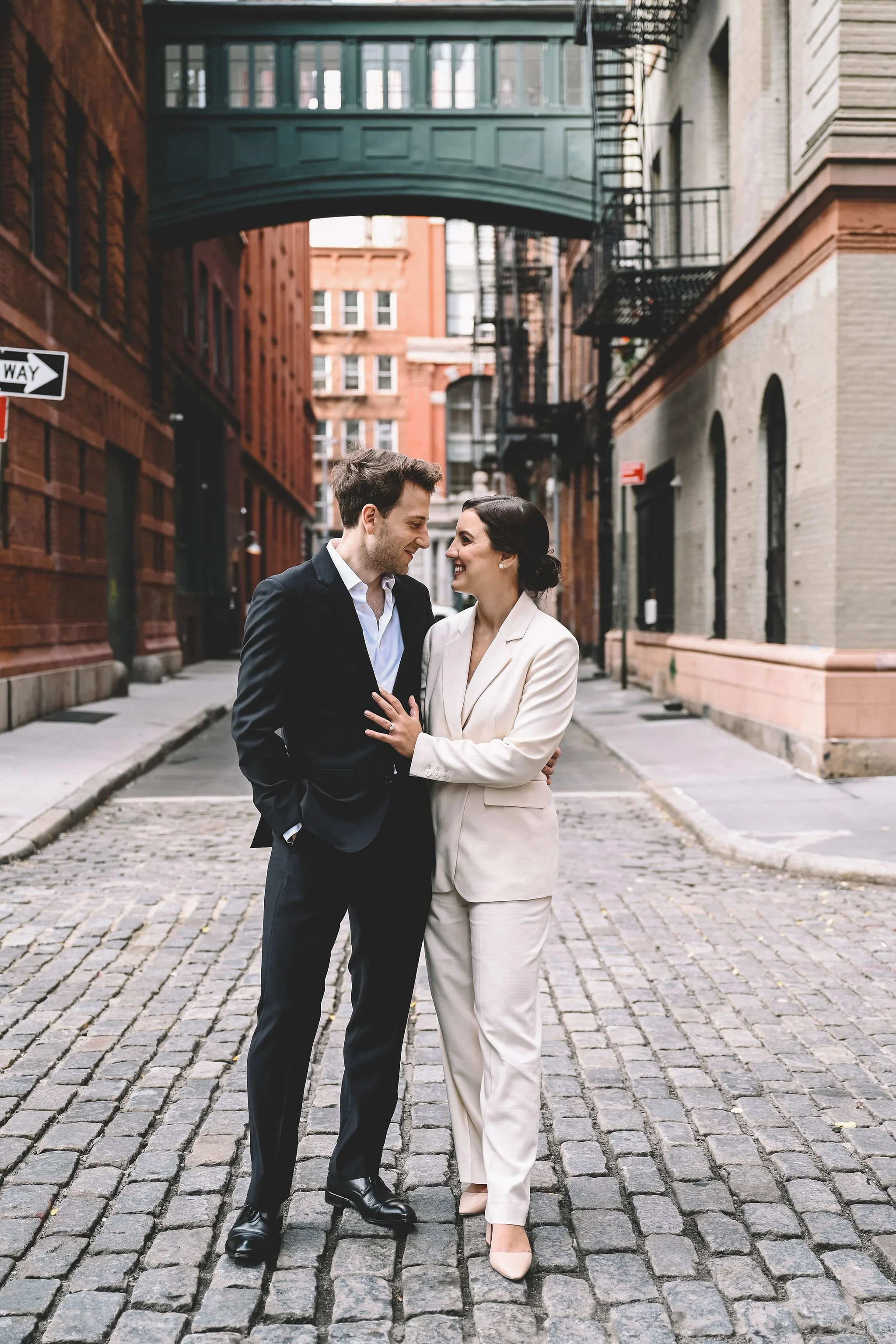 A couple dressed in business attire standing close together and smiling on a cobblestone street in an urban area.