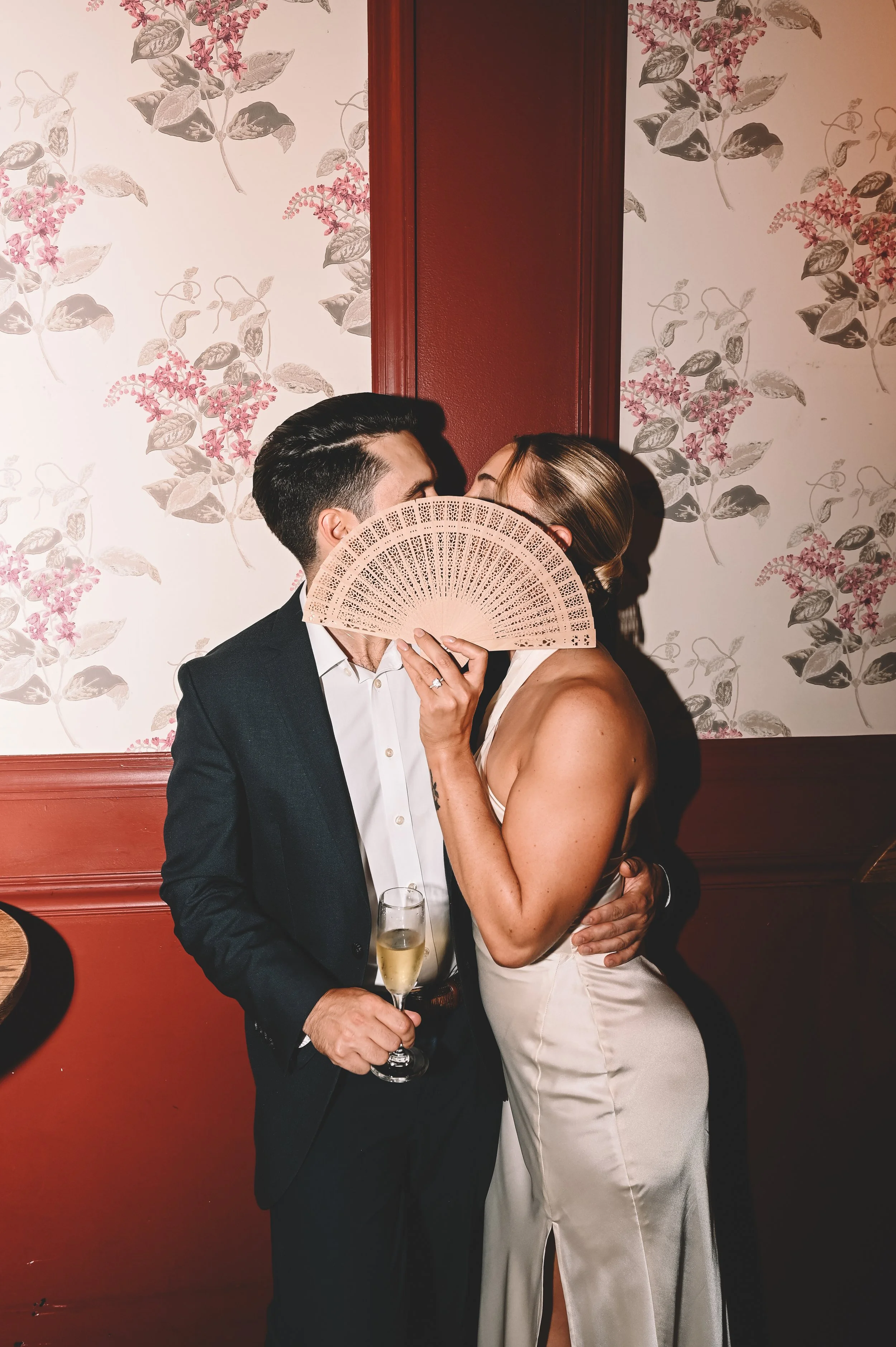 A couple dressed in formal attire sharing an intimate moment at a wedding or event. The man is in a suit holding a champagne glass, and the woman in a satin dress is holding a lace fan, partially covering their faces. They are standing against a floral patterned wallpaper wall.