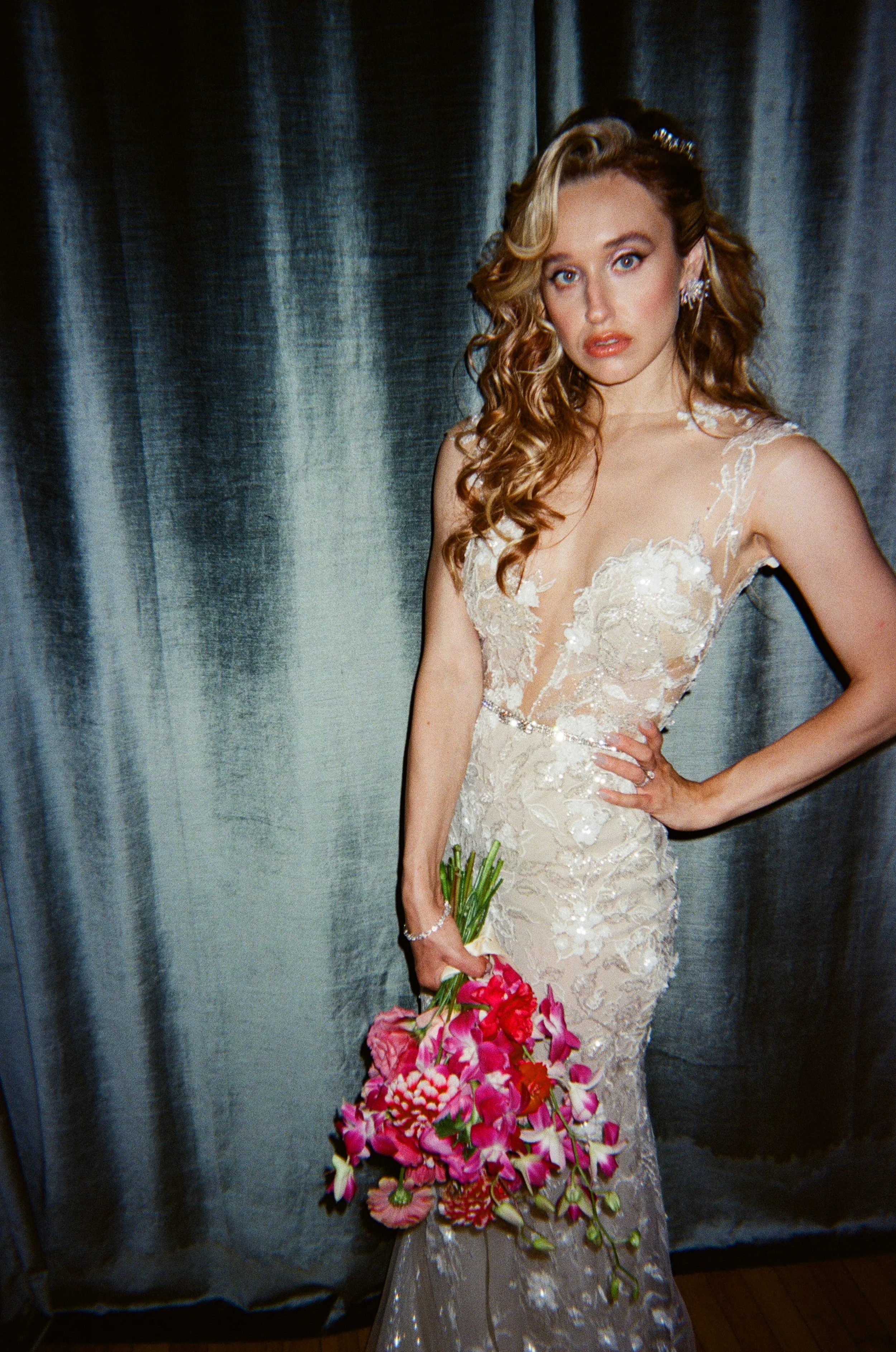 A woman in a wedding dress holding a bouquet of pink and red flowers, standing in front of a gray curtain.