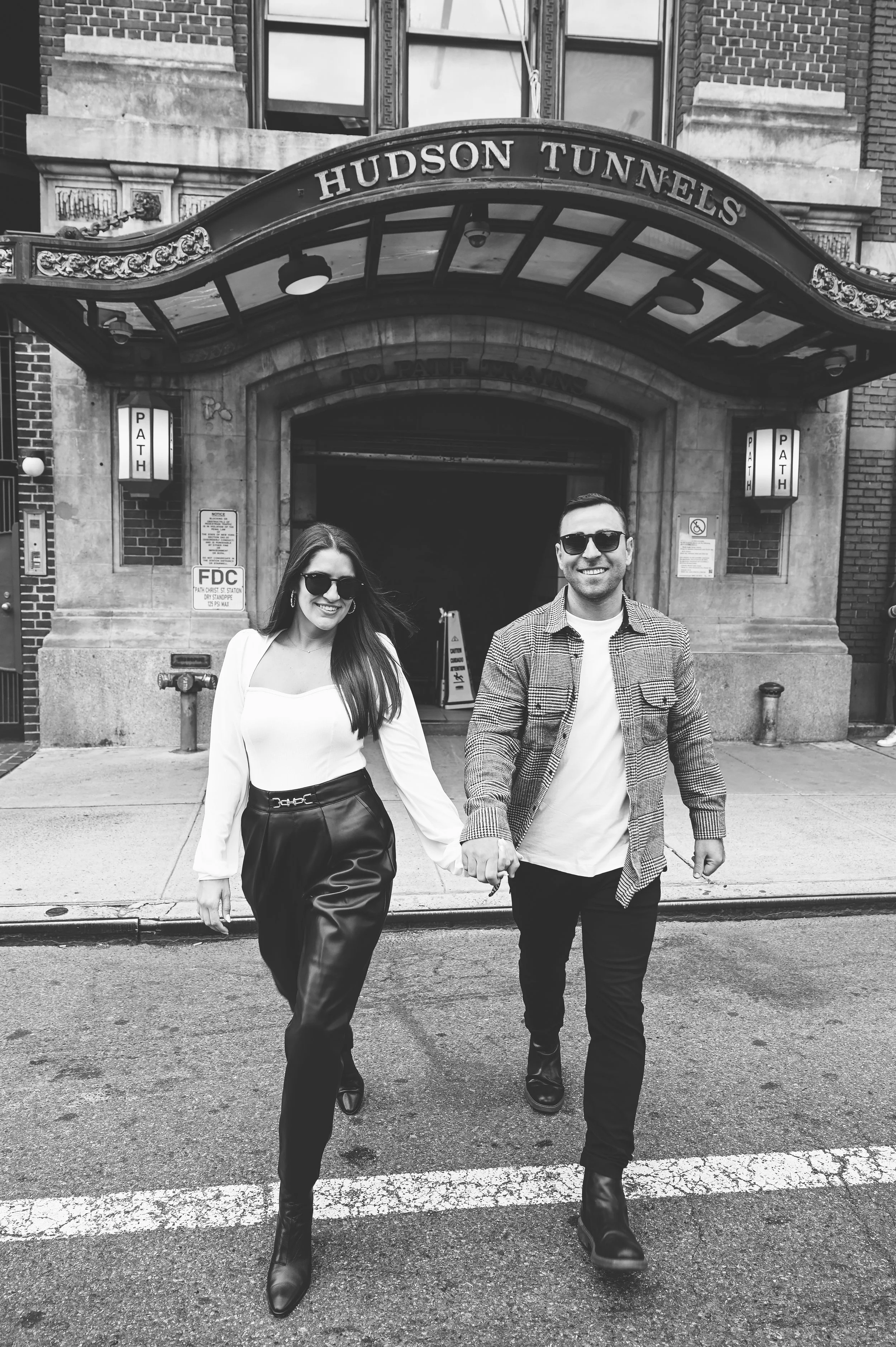 A smiling couple holding hands and walking out of the Hudson Tunnels entrance, dressed stylishly in casual outfits, with the tunnel's sign visible above.