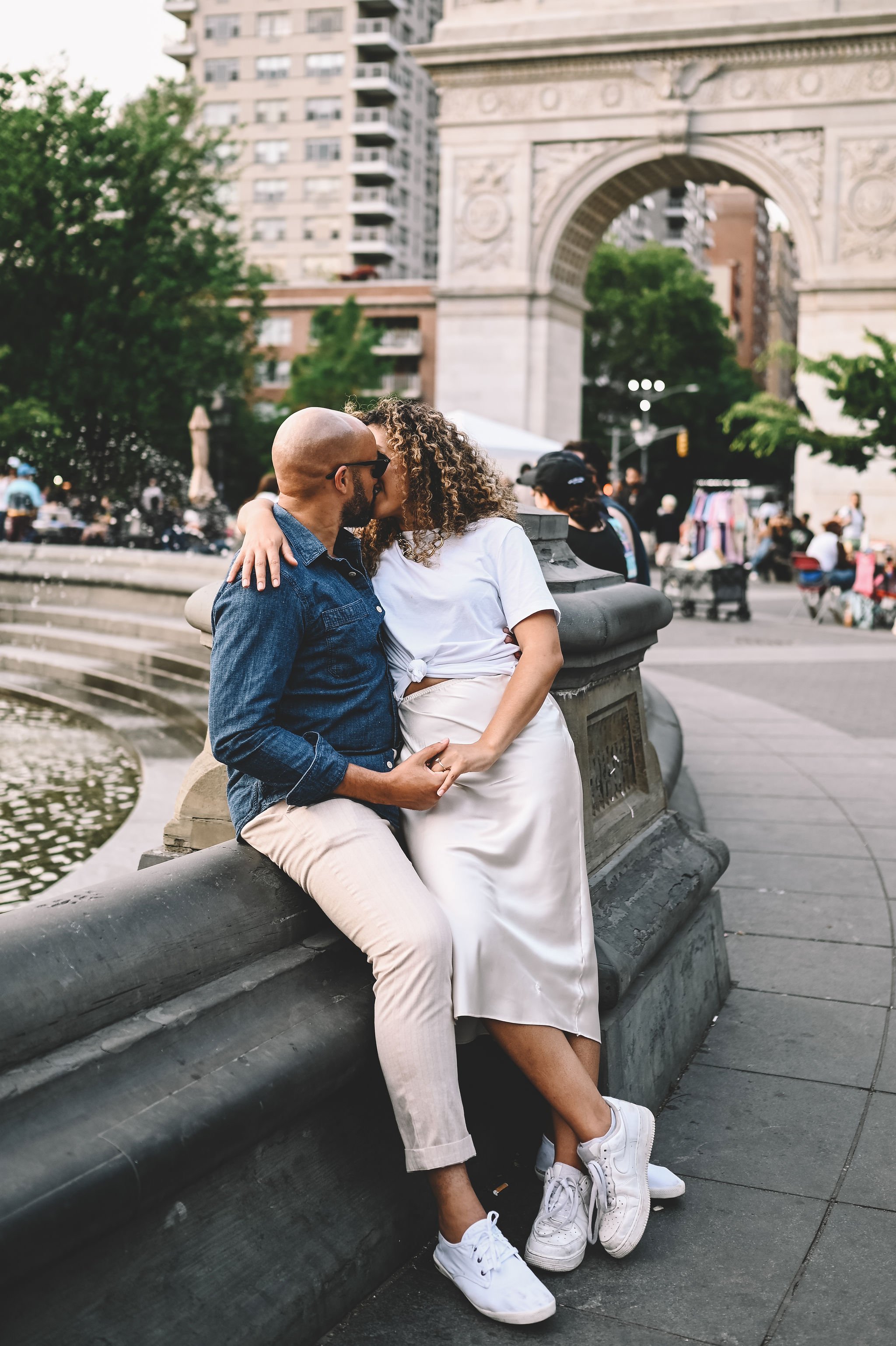 A couple kissing on a stone ledge in a city park with an arch monument in the background.