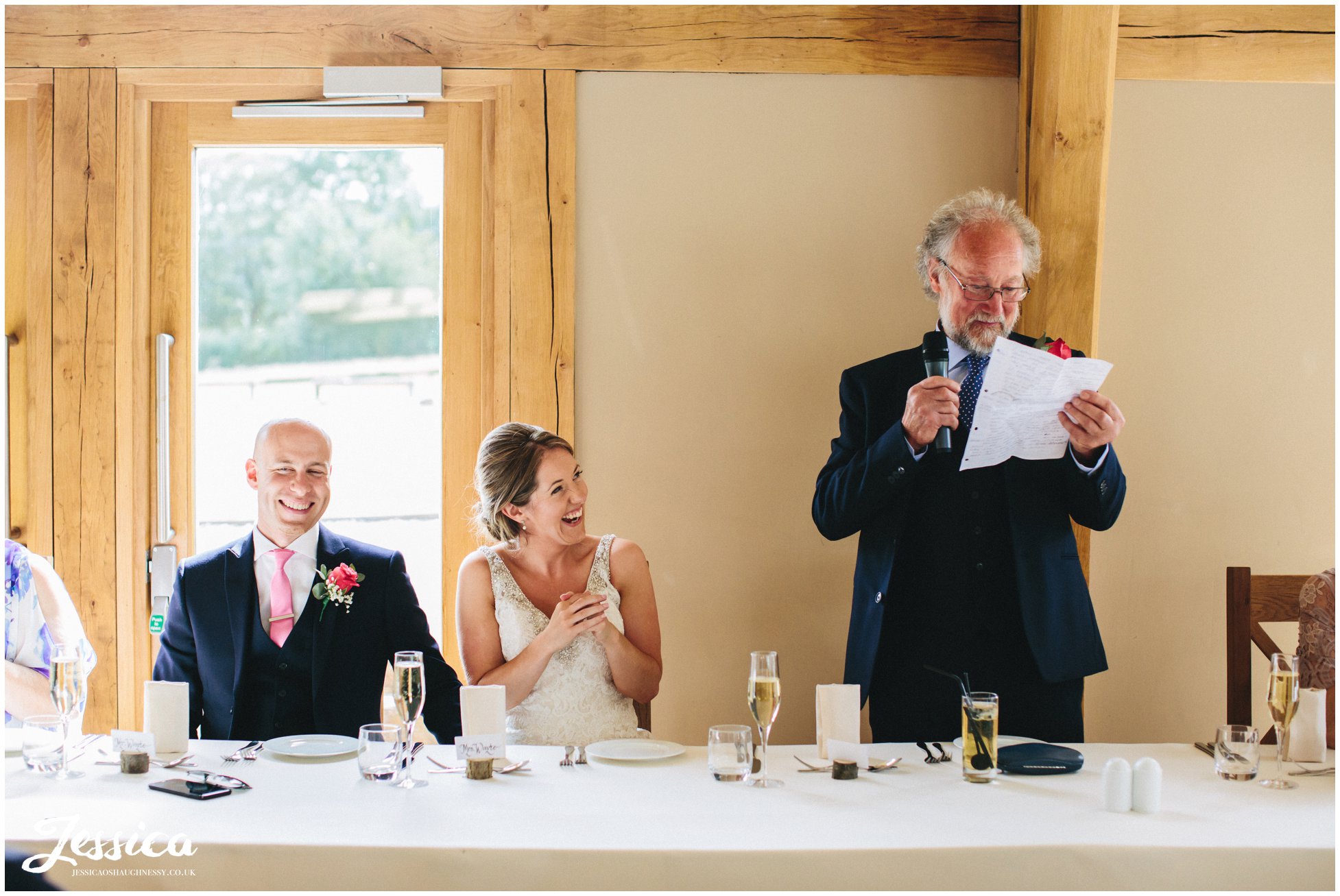 father of the bride stands and gives speech whilst the bride laughs