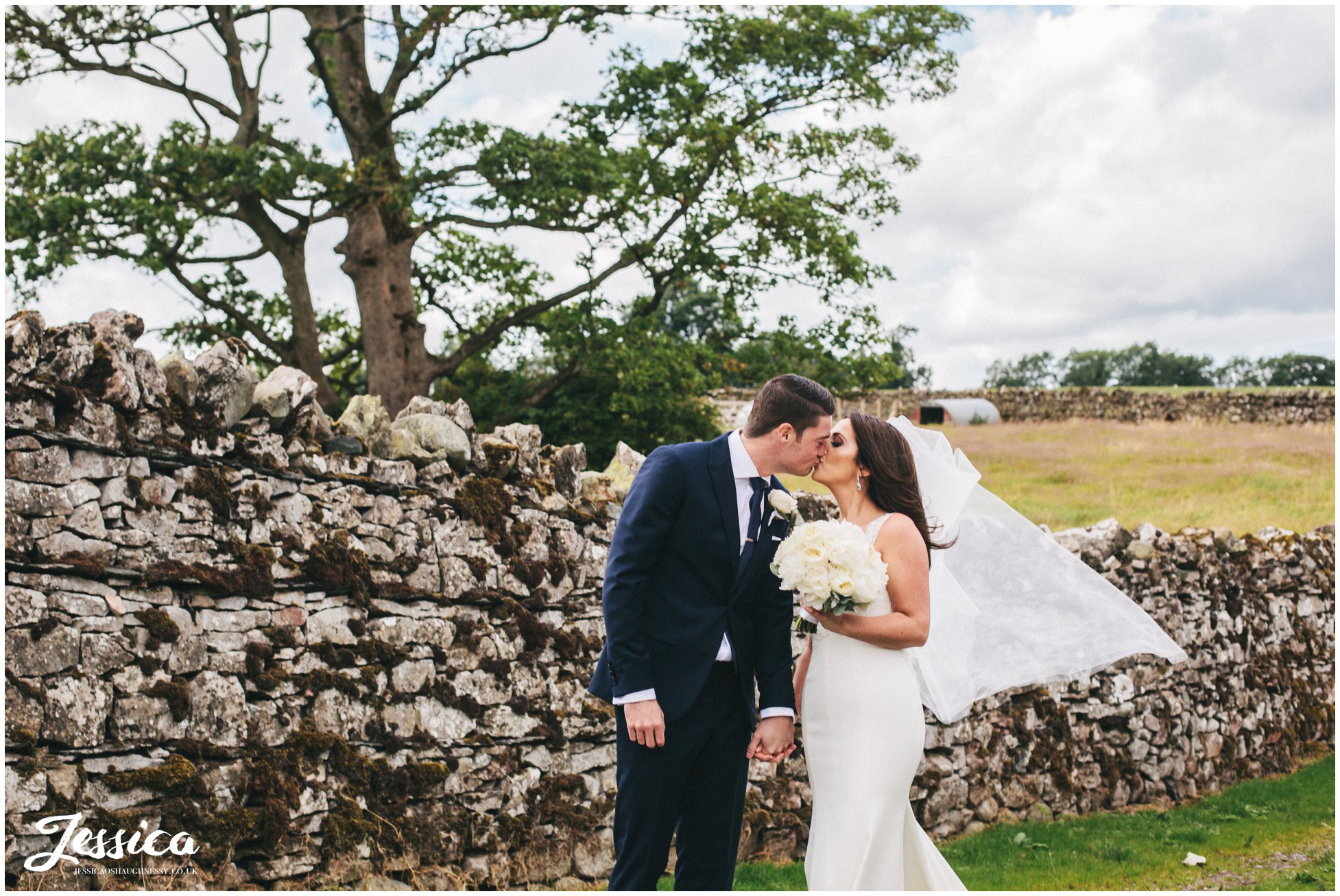 newly wed's share kiss in front of stone wall after their wedding at askham hall in the lake district