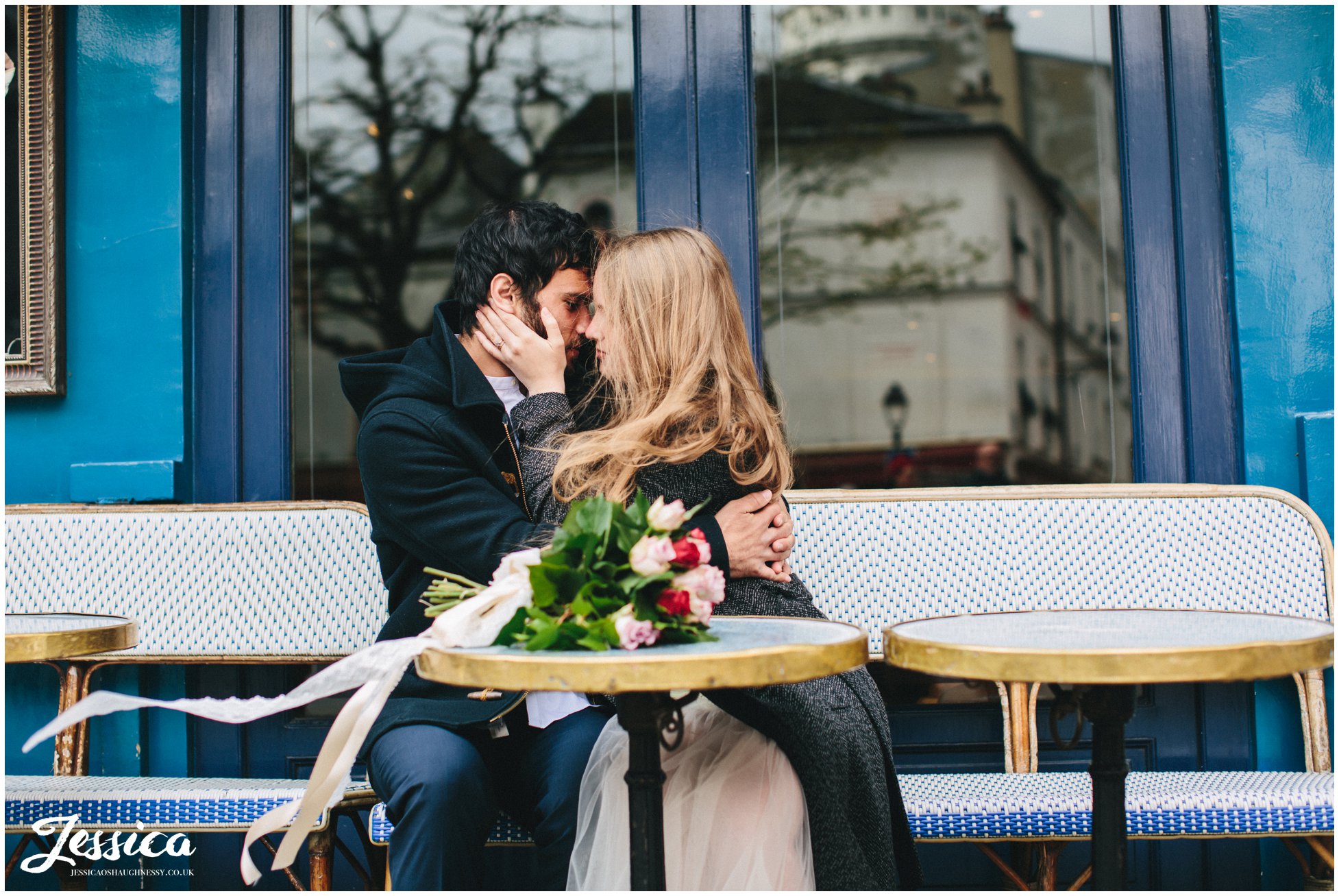 wind blows brides hair as she kisses her groom sitting in a cafe in mont marte