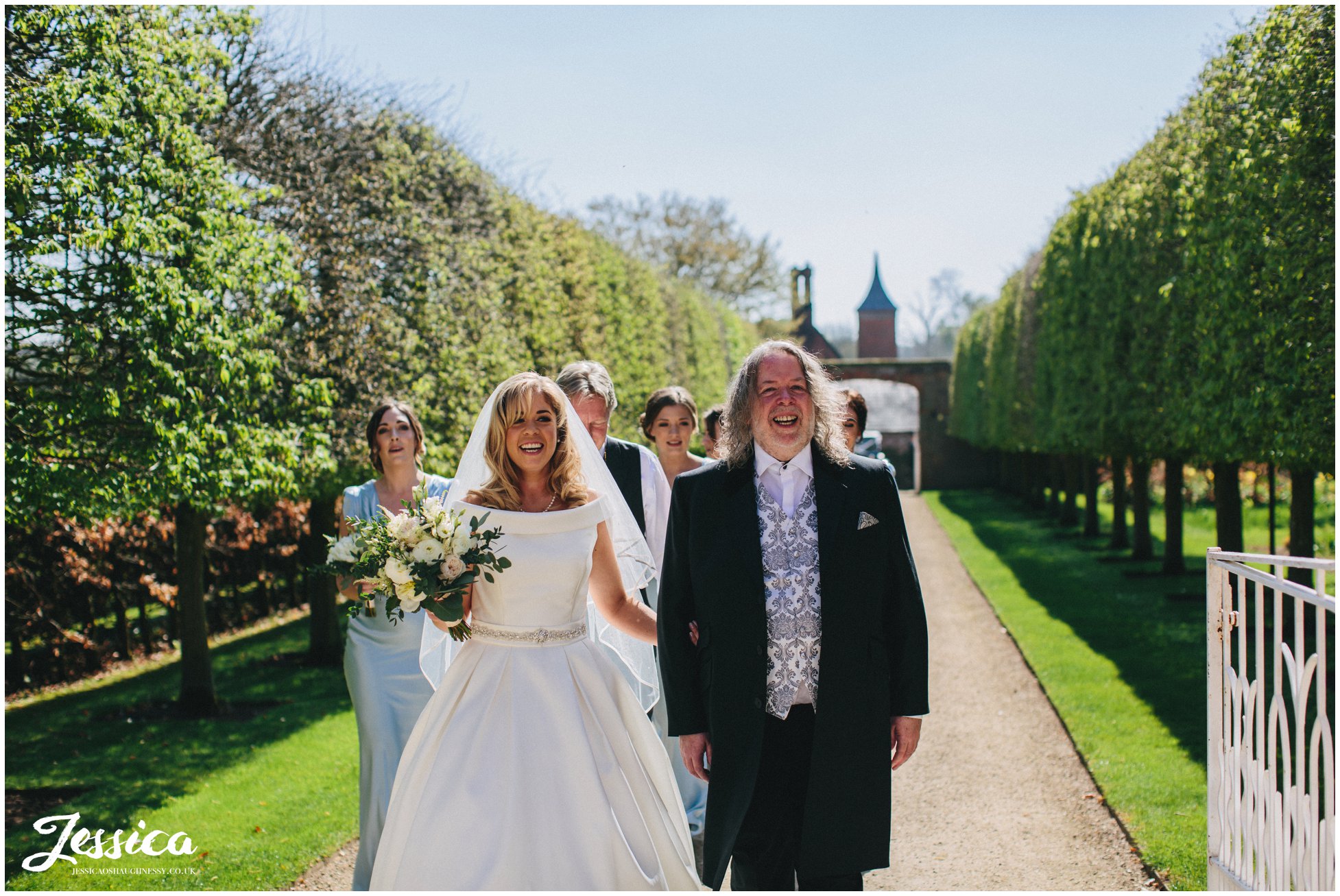 bride & her father walk down path at Combermere Abbey to the ceremony in the glasshouse