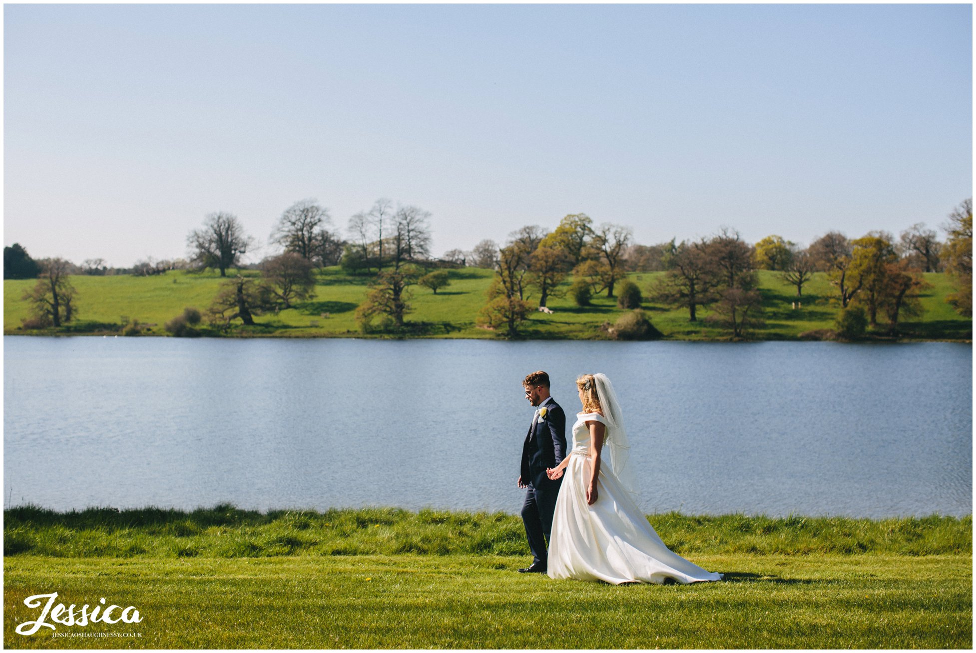 couple walk holding hands along side the lake on a sunny day