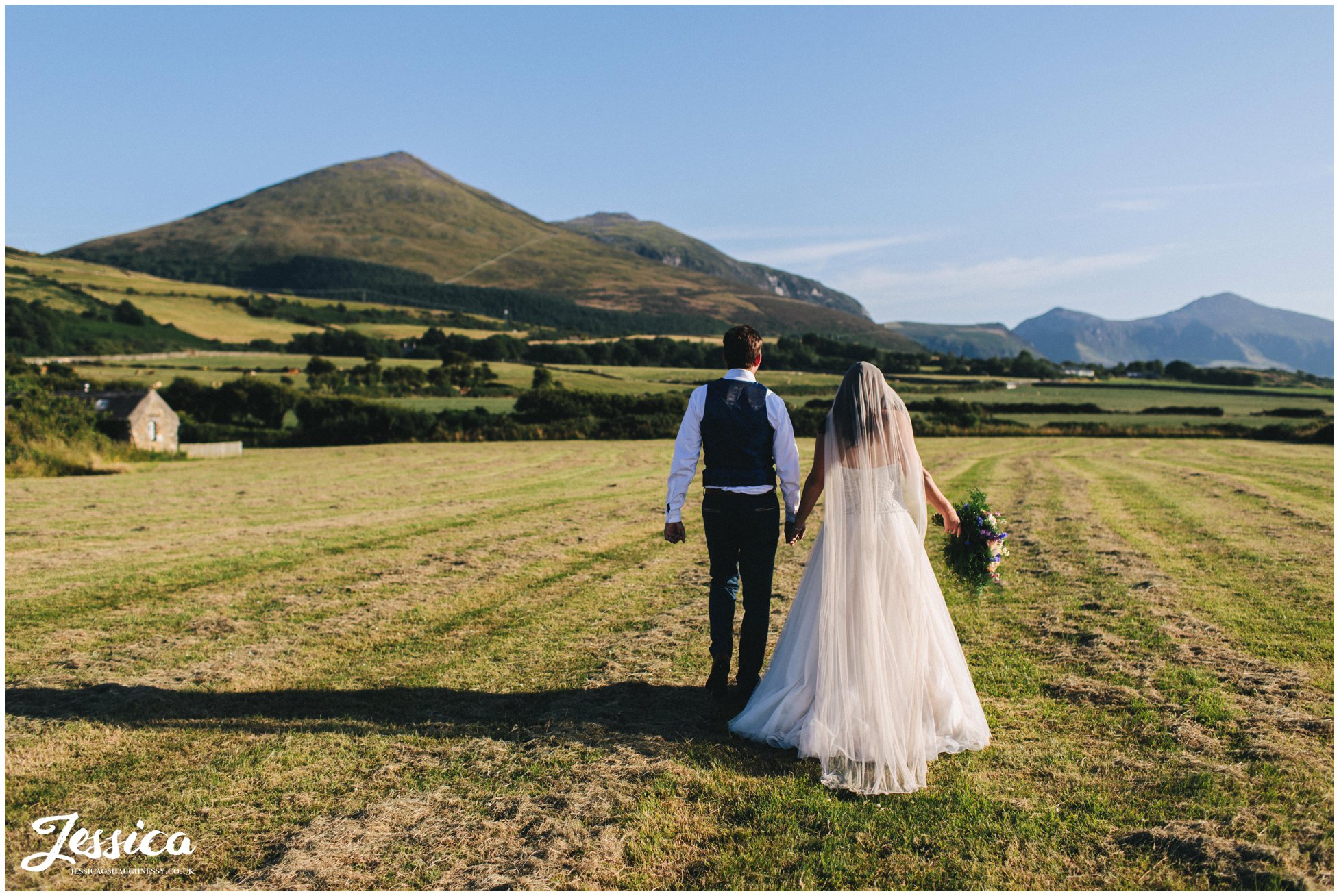 husband and wife walk hand in hand in front of the Welsh hills