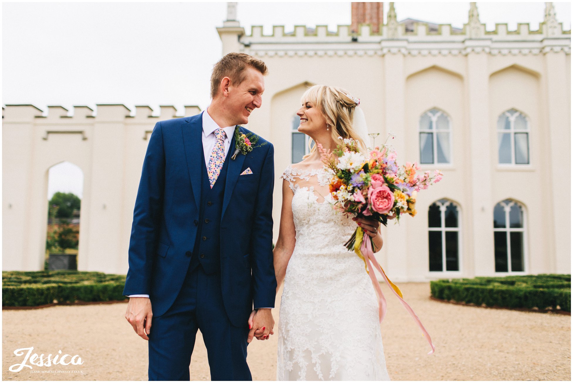 husband and wife hold hands walking in front of combermere abbey