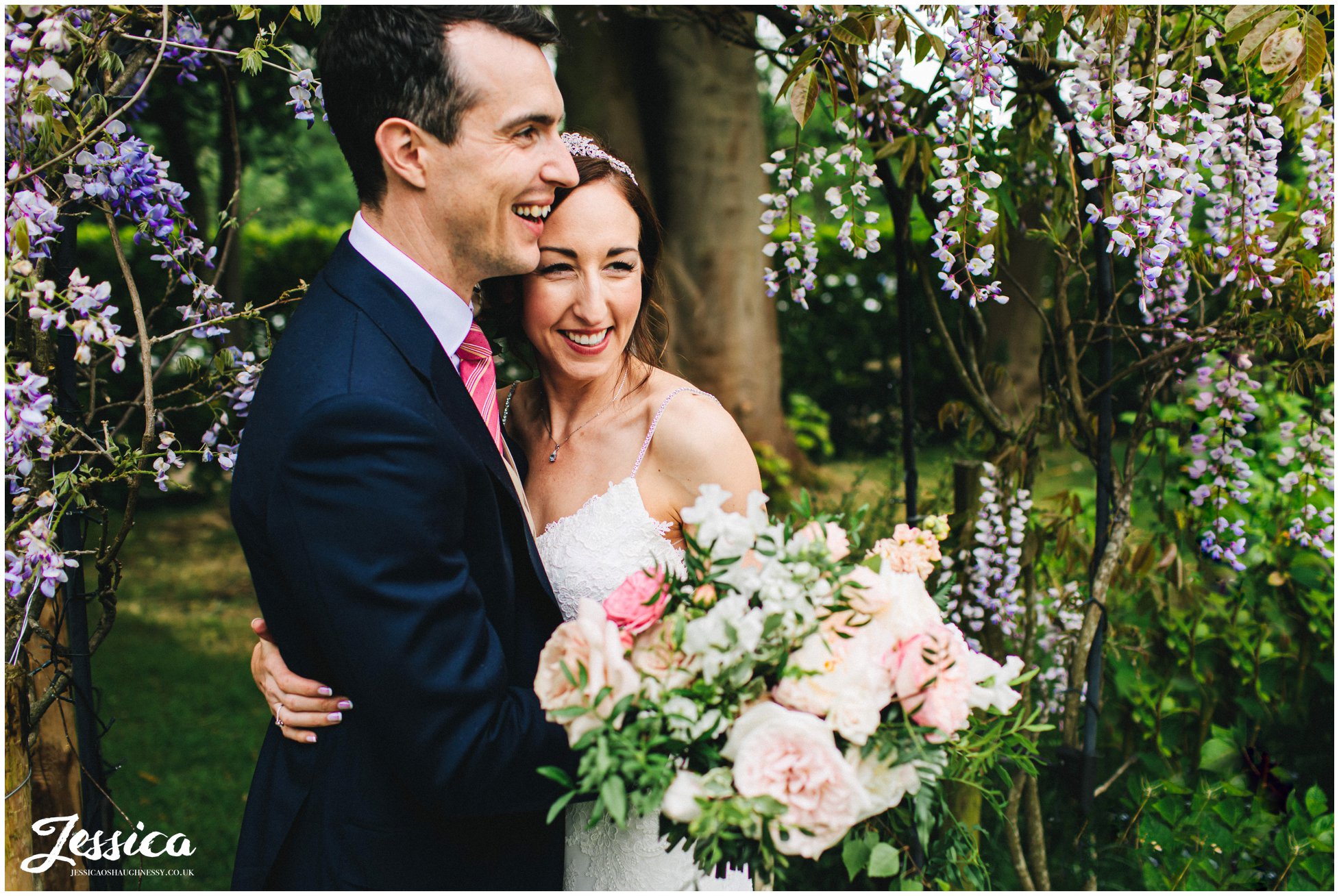 bride &amp; groom stand under wisteria in the gardens