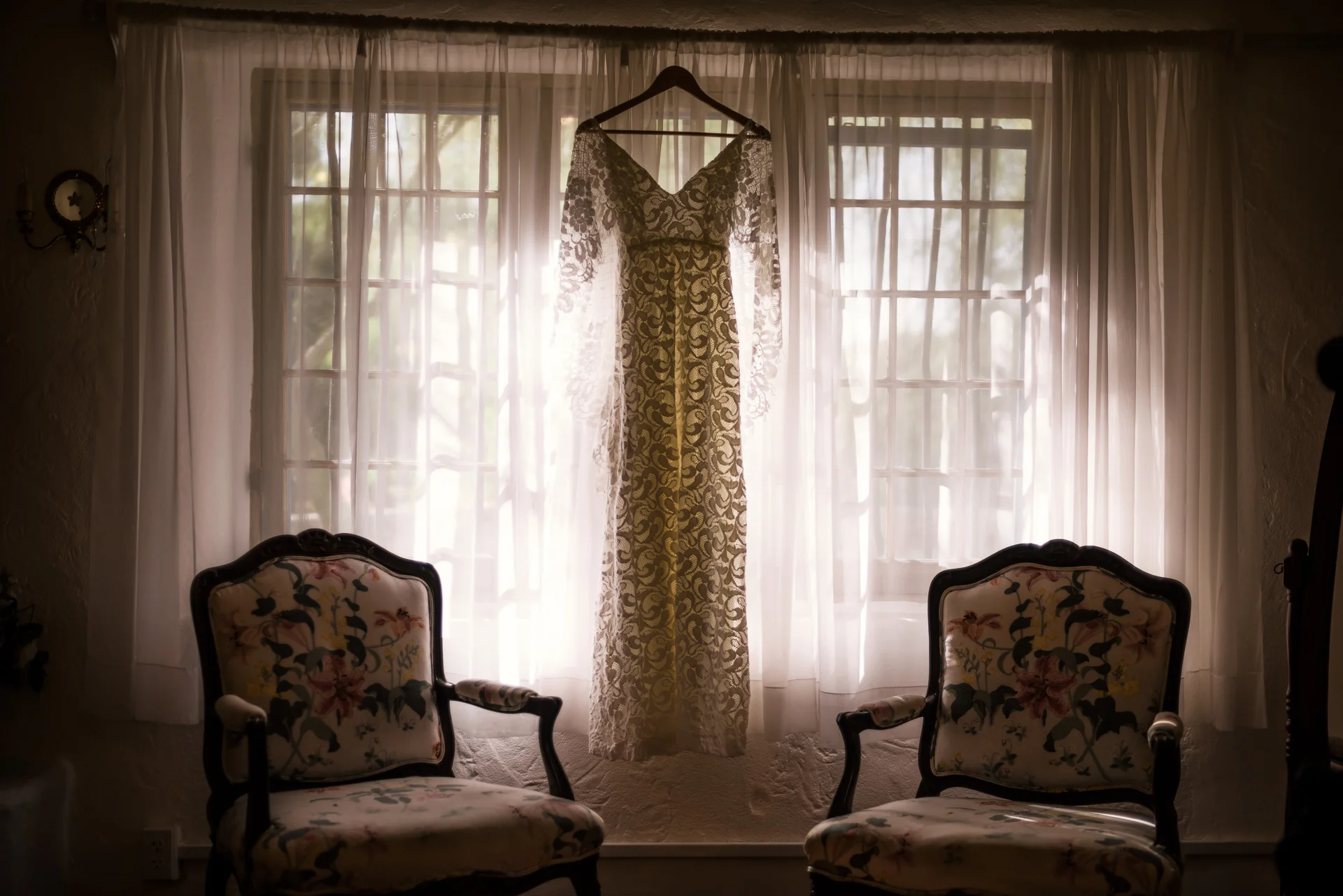 Brides wedding dress hangs backlit by window light between two chairs captured by photographer during romantic wedding at the historic Leo Carrillo Ranch in Carlsbad California