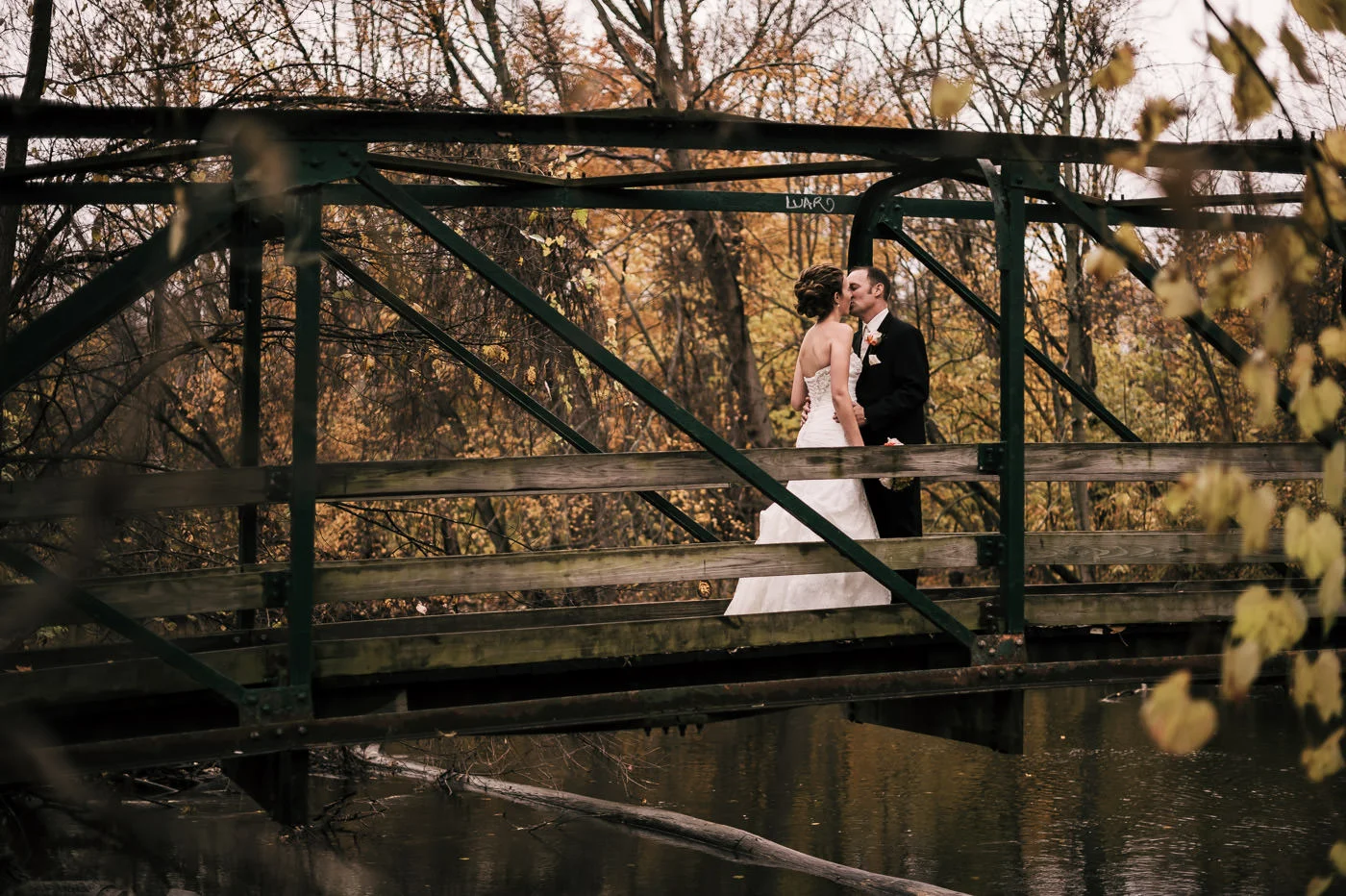 Bride and groom have a romantic kiss on an old wood and steel bridge over a river among the stunning colors of fall in Michigan.