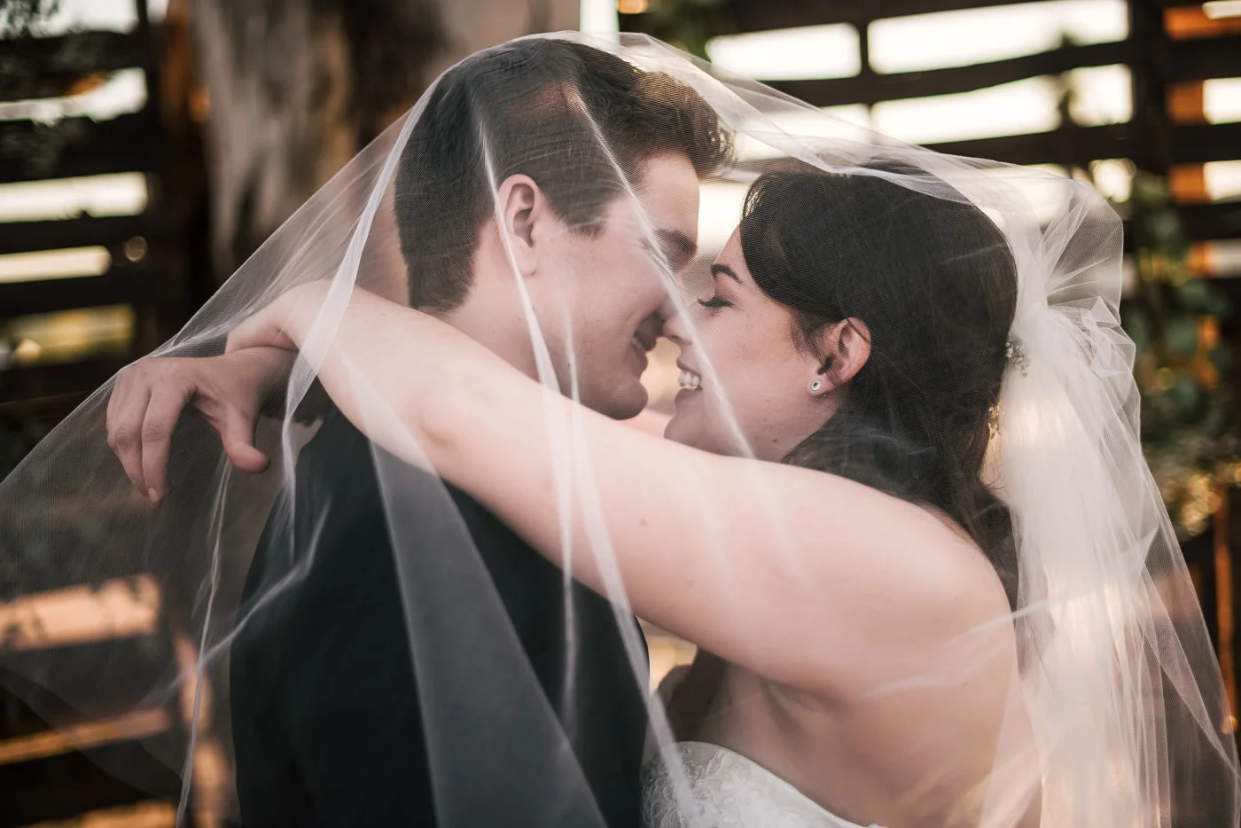 Bride and groom kiss under the wedding veil in front of a pallet wall at their Temecula marriage ceremony.