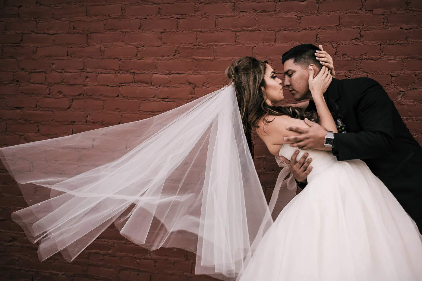 Groom dips bride romantically in front of a red brick wall as her veil blows in the wind.