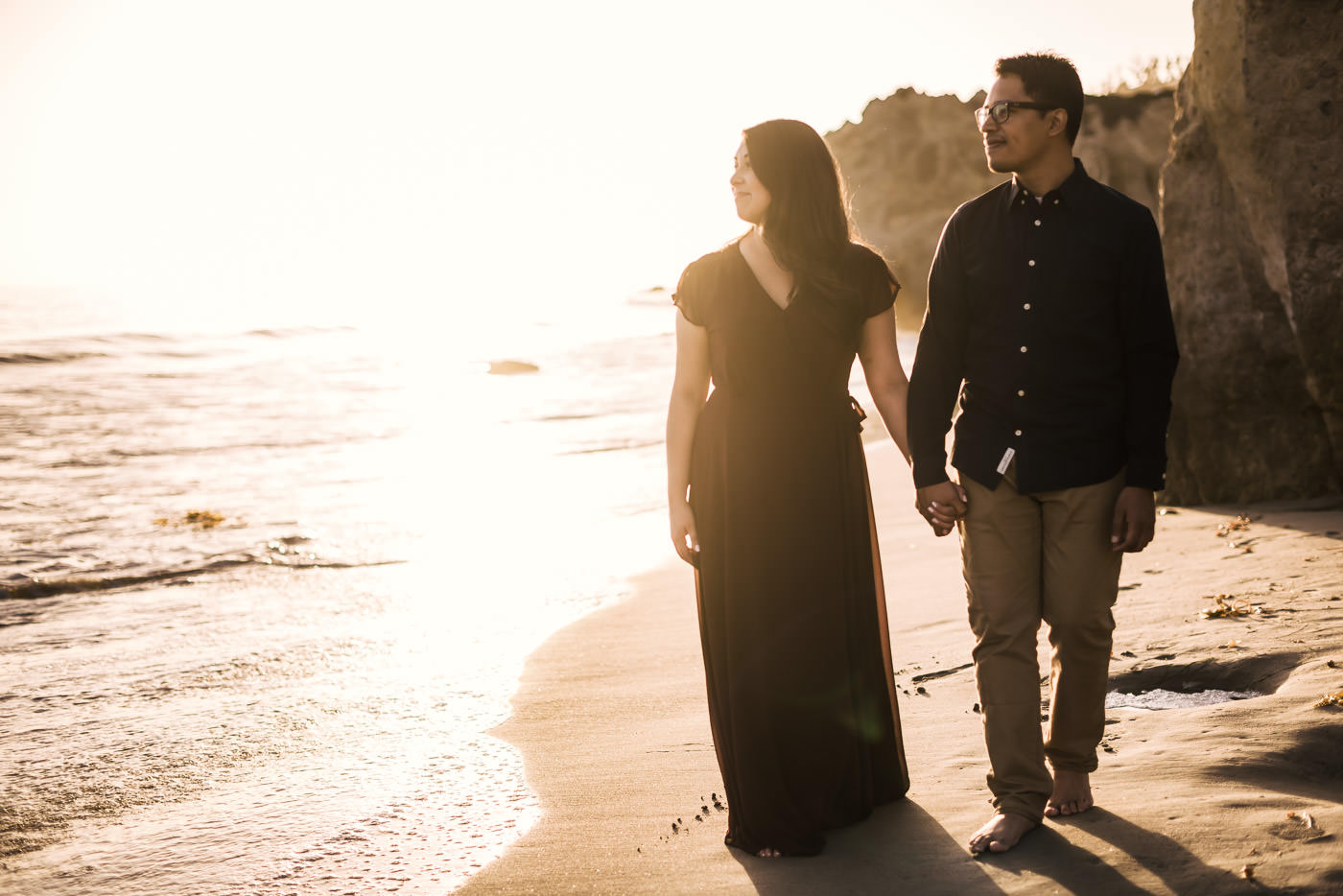 Couple walks hand in hand across El Matador State Beach as they stare off into the ocean and the setting sun in Malibu California