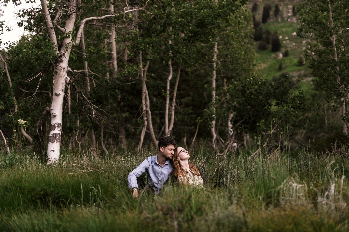 Engaged couple sits in the tall grass of a meadow backed by birch trees in the mountains of Sequoia national park for their engagement shoot