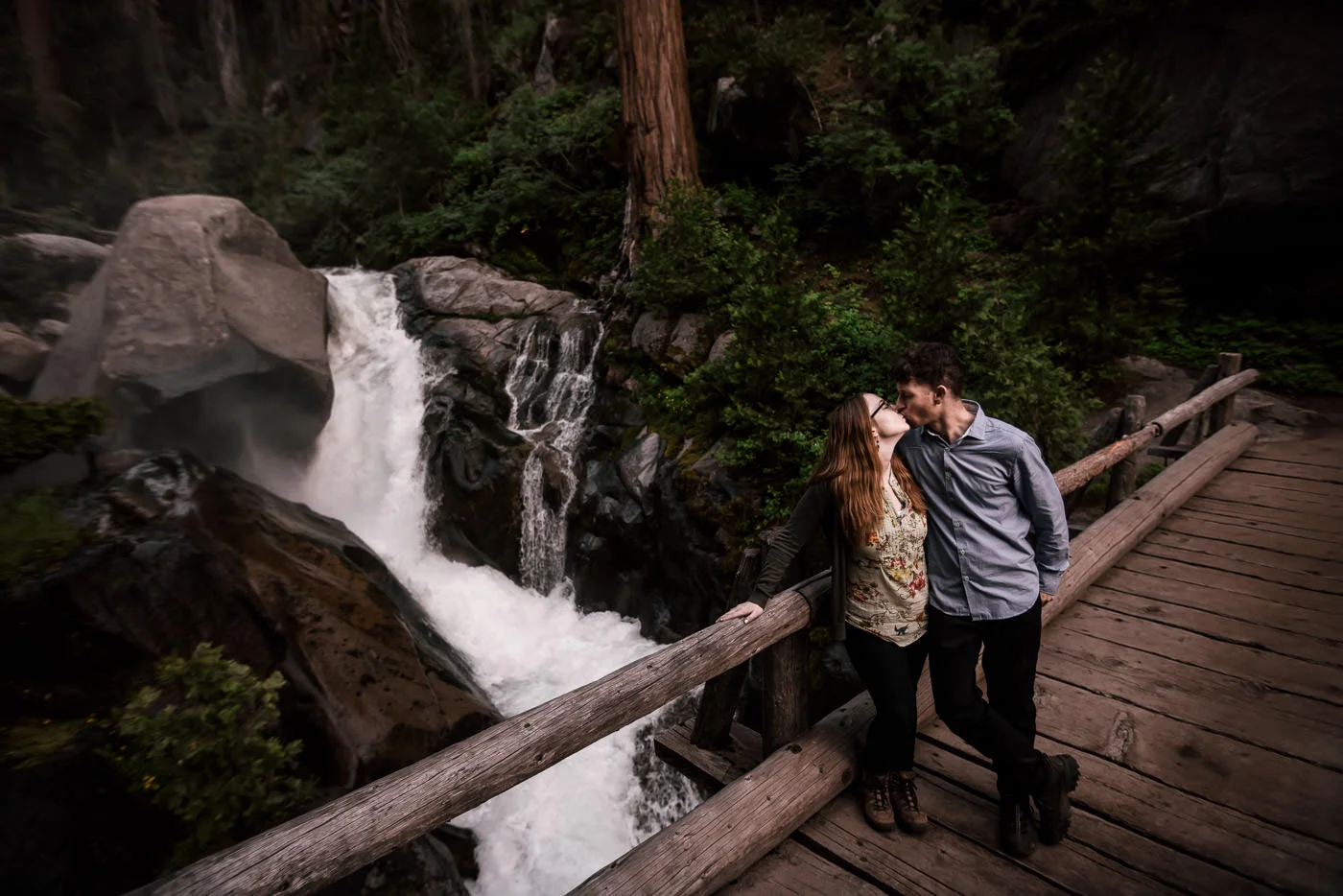 future bride and groom share a kiss on a bridge in front of a raging waterfall during their engagement session in seqouia national park
