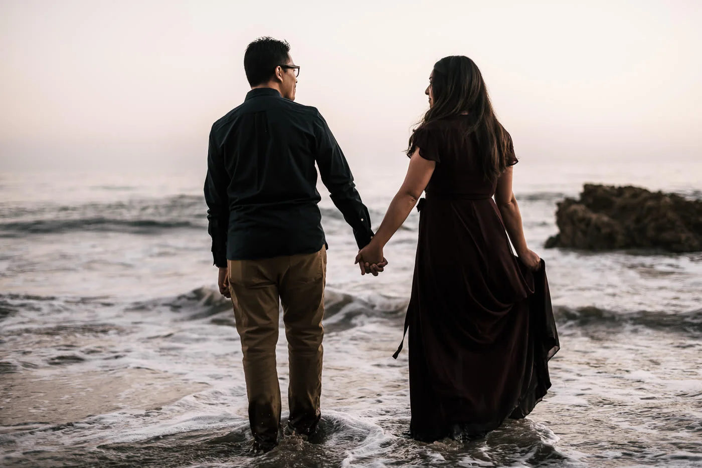 Sweet couple walks into the ocran holding each others hands tightly at their El Matador State Beach engagement session in Malibu California.