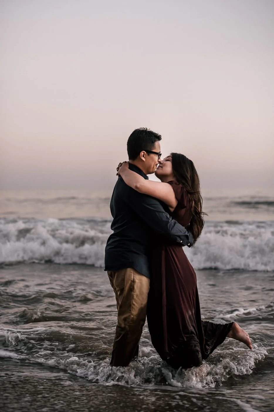 Couple walks out into the ocean at sunset and has a romantic photo session for their engagement in Malibu California. The bride to be pops her leg as they kiss for a classic photograph.