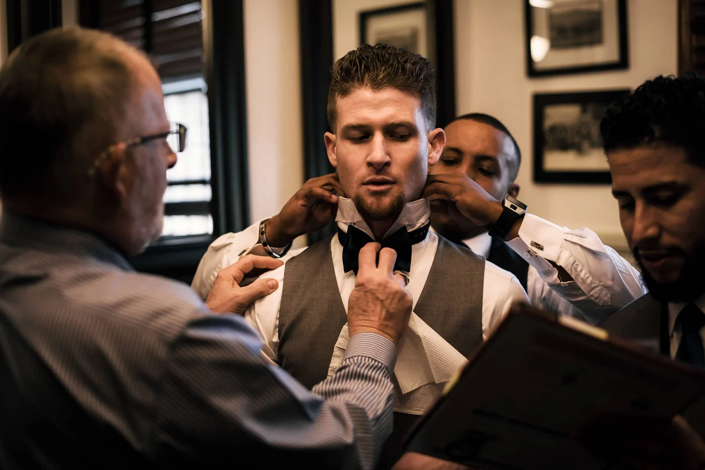 Groomsmen help the groom get ready while he dictates his vows to the best man.