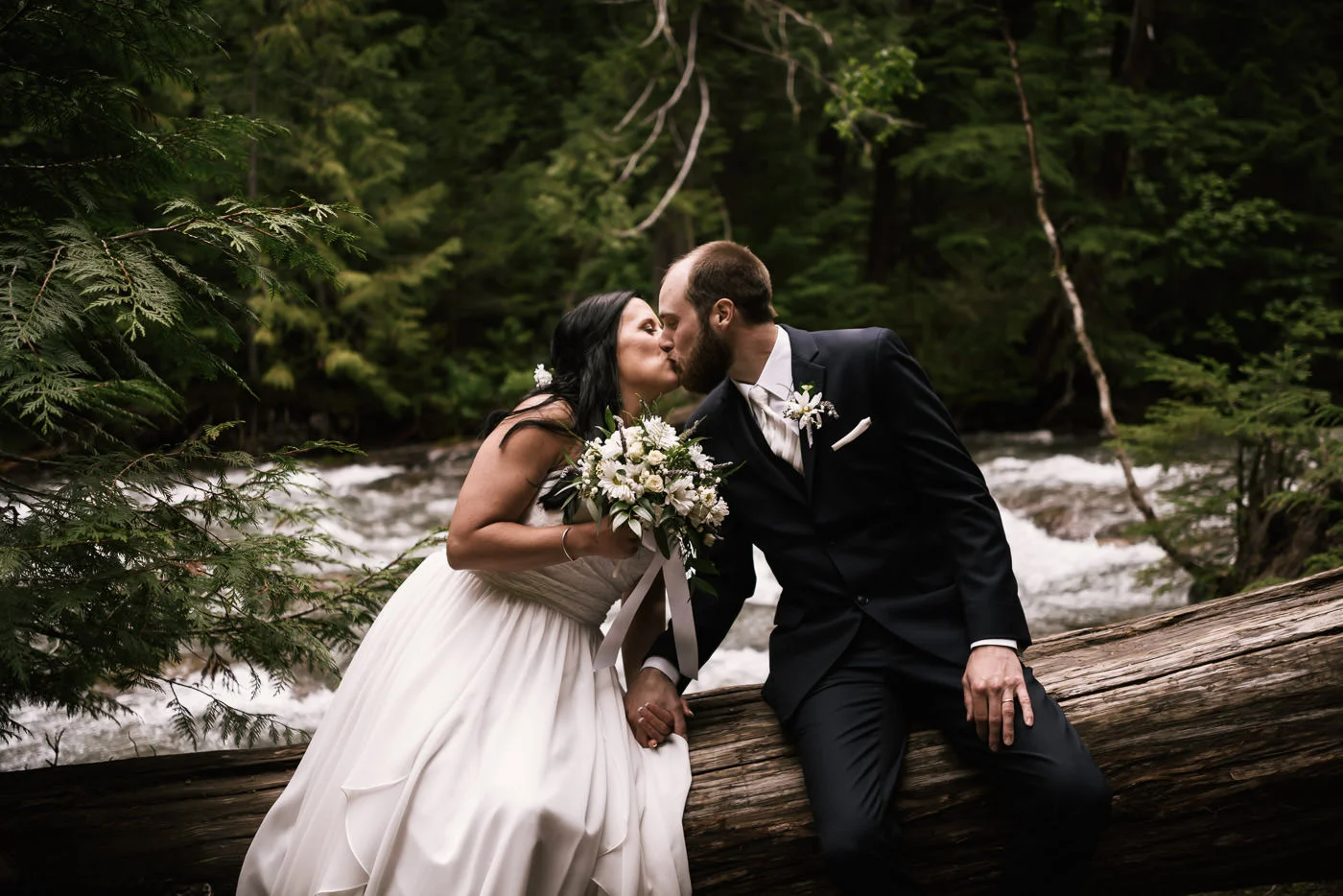 Couple in love sits on a fallen cedar tree and kiss in front of a raging river.
