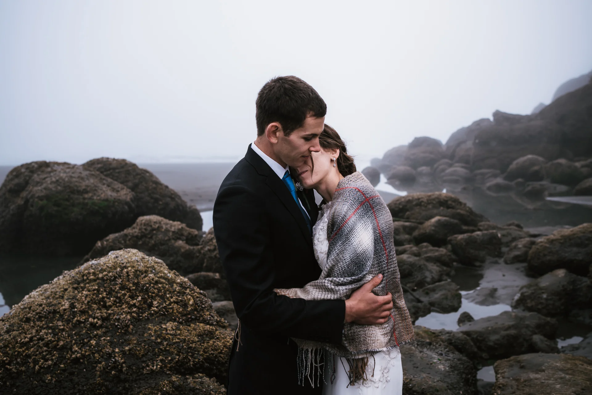 Among the barnacle covered rocks and thick fog, a couple holds each other tightly and shares a quiet moment.
