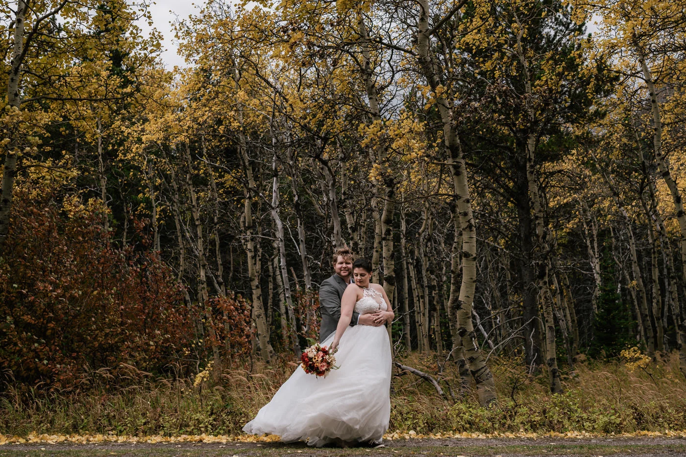 Groom wraps his arm around his bride in front of a birch forest with fall color in Many Glacier.