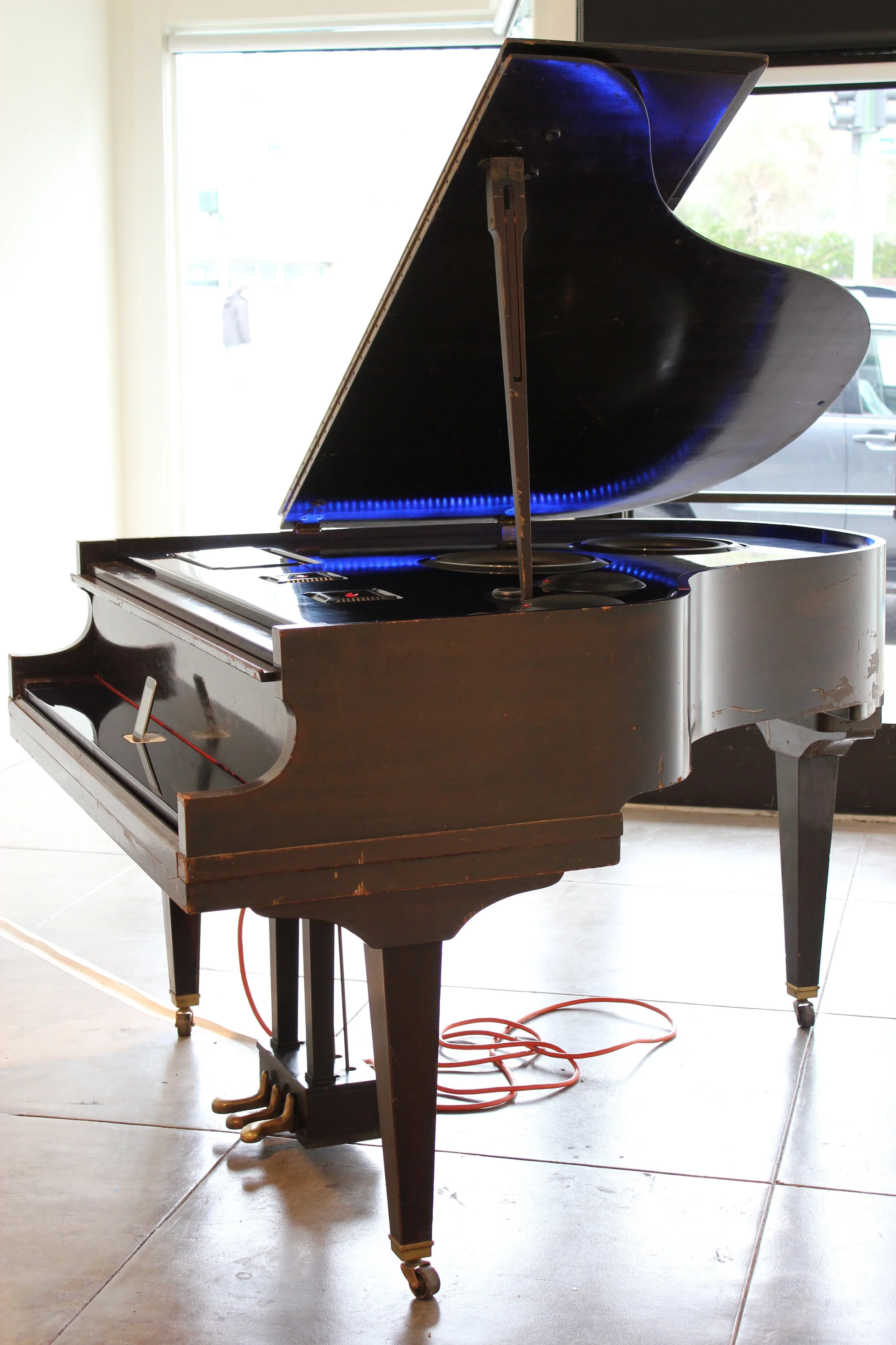An upright black grand piano with its lid open, Fosgate speakers replacing the strings and hammers inside, situated indoors on a tiled floor.
