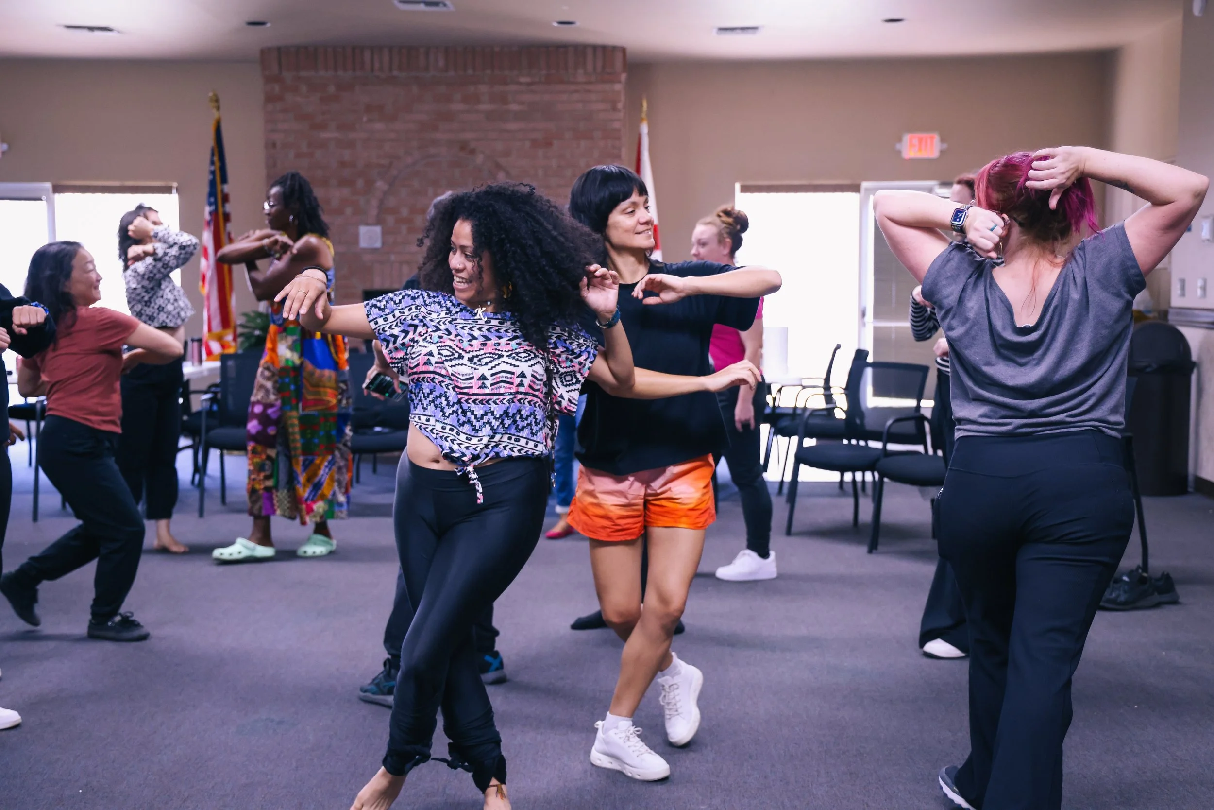A group of people of different genders, ages, and ethnicities are dancing, with arms raised, around a room
