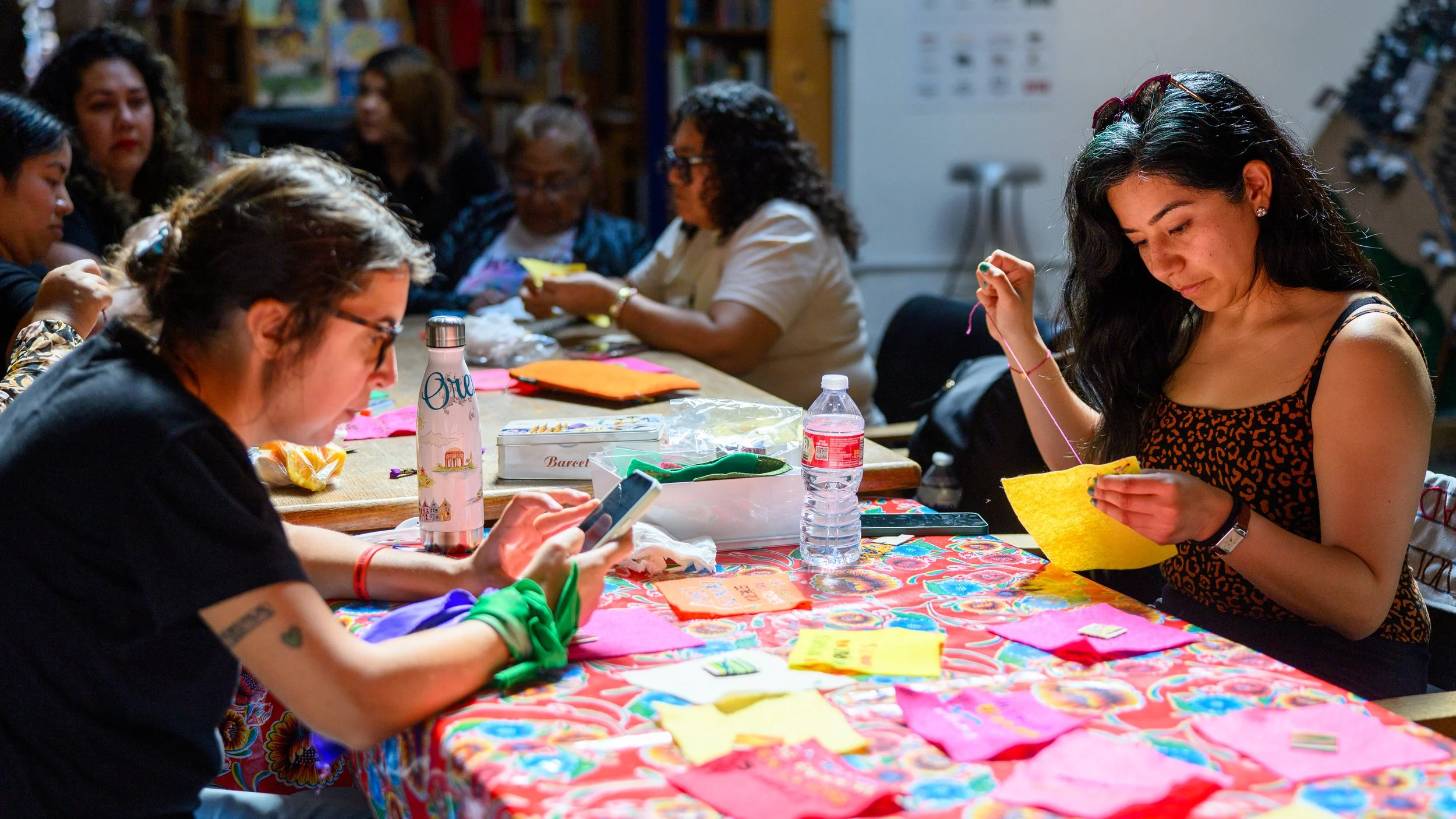 A group of women sit around a brightly colored table, embroidering colorful pieces of felt.