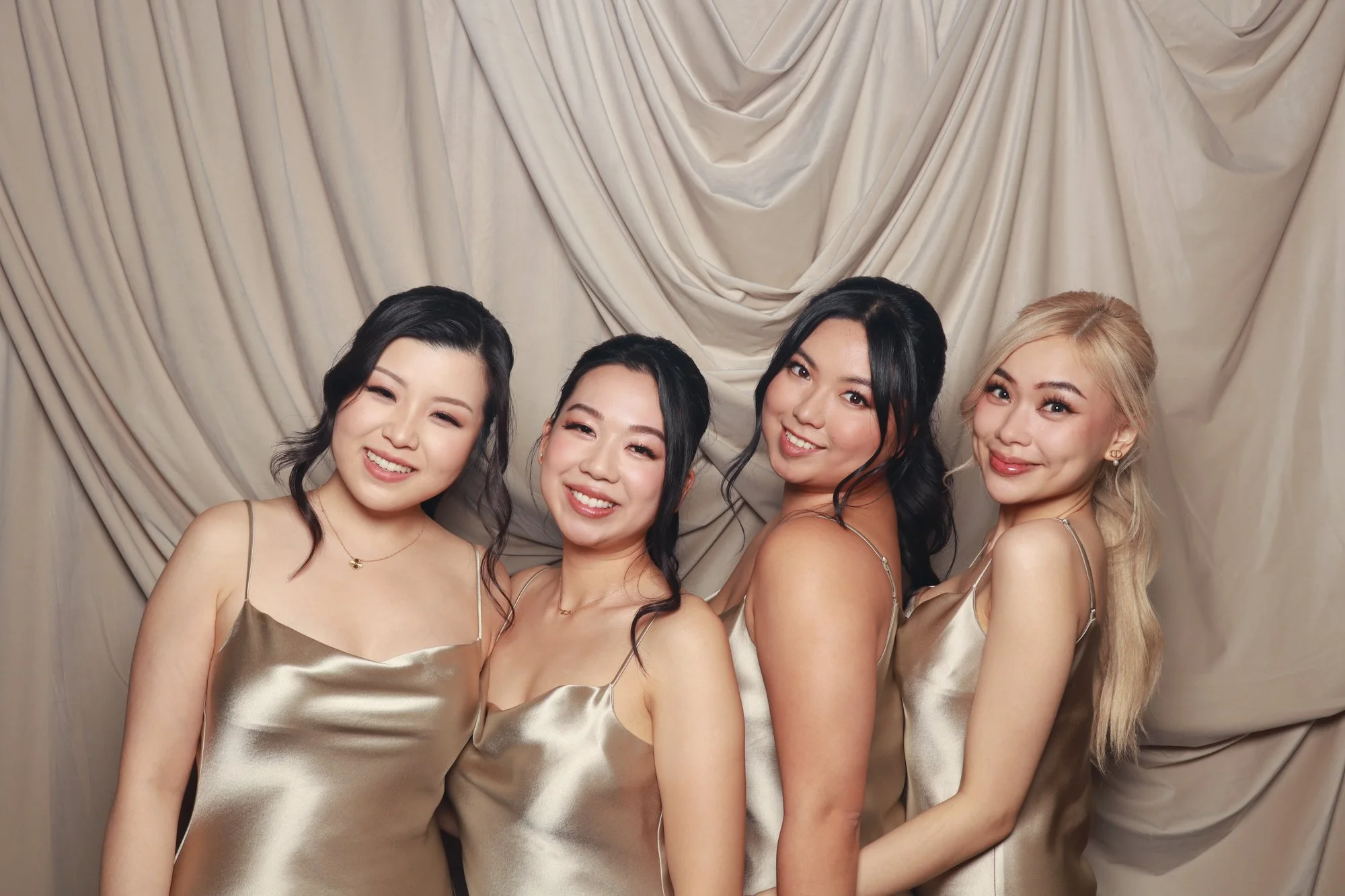Four wedding guests in tonal satin dresses posing against a warm neutral velvet drape photo booth backdrop.