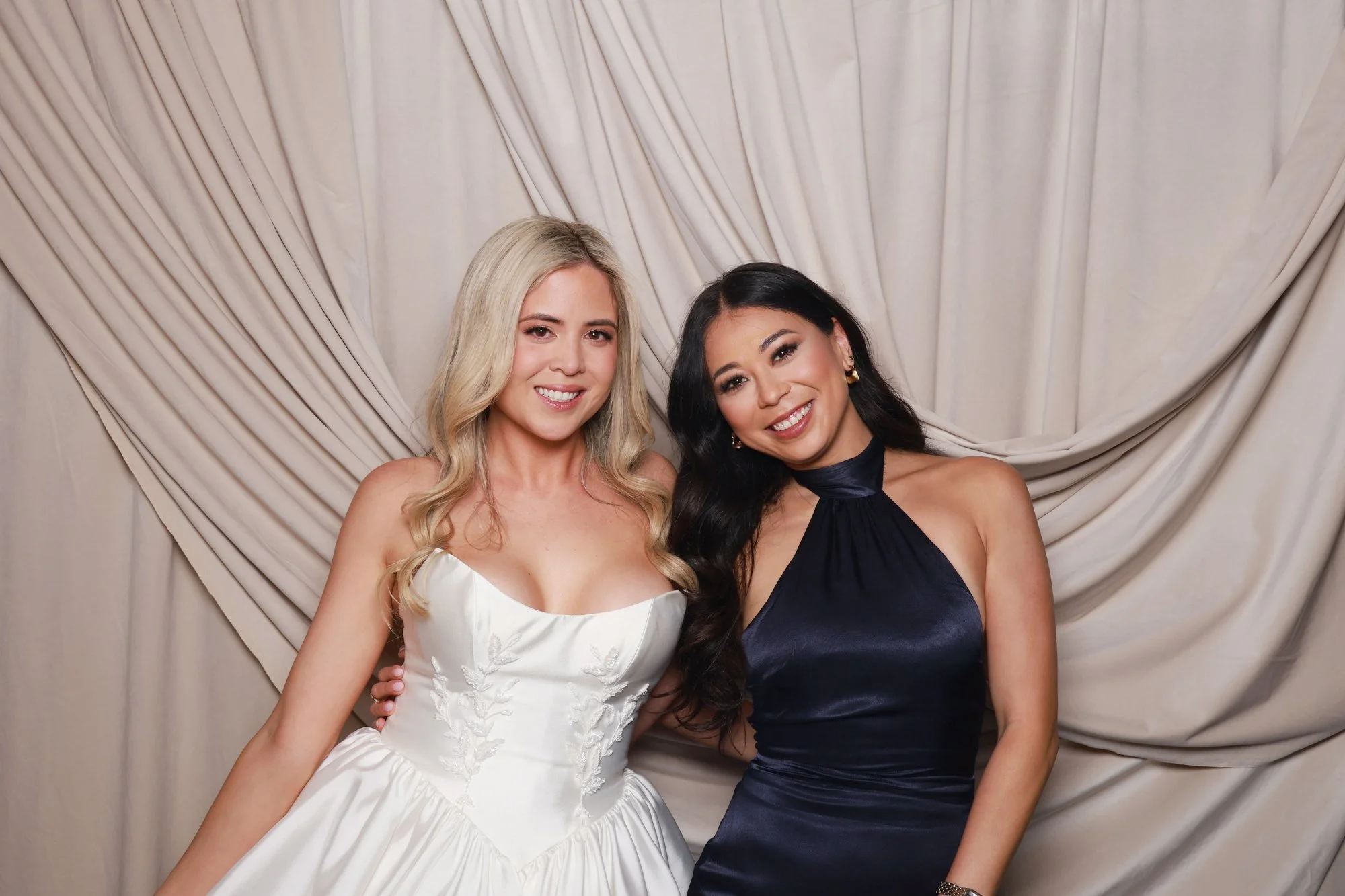 Bride with wedding guest posing against the soft layered folds of the Golden Hour Velvet Drape photo booth backdrop.