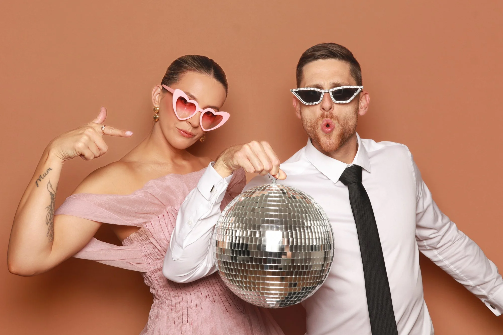 Wedding guests posing with a disco ball and heart-shaped sunglasses in a Photo Corner photo booth.