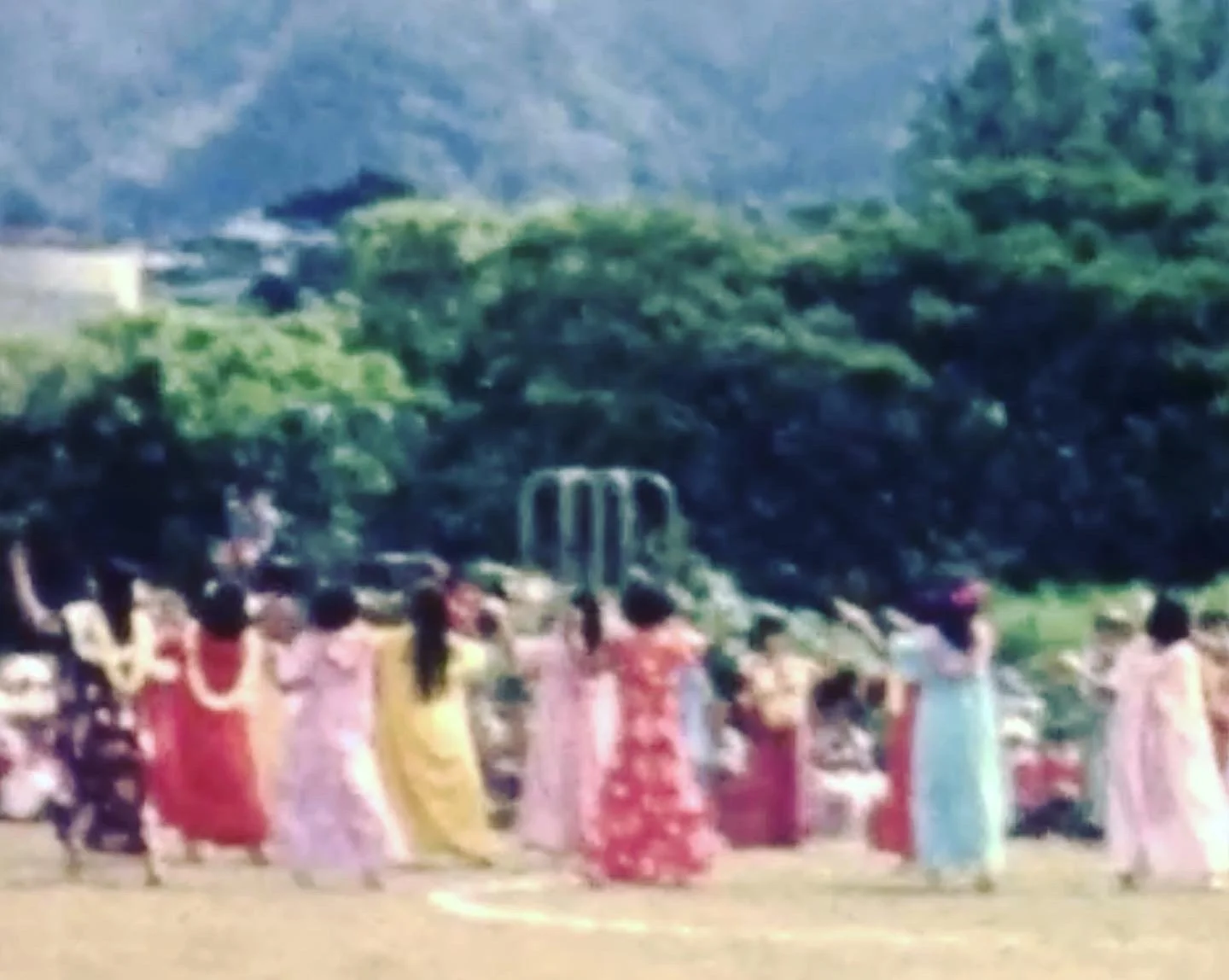 Still image from an old reel of Hawaii elementary school Lei Day pagent in the 70's. A group of women in colorful mu‘umu‘u wearing lei dance in a field with trees, & mountain in background