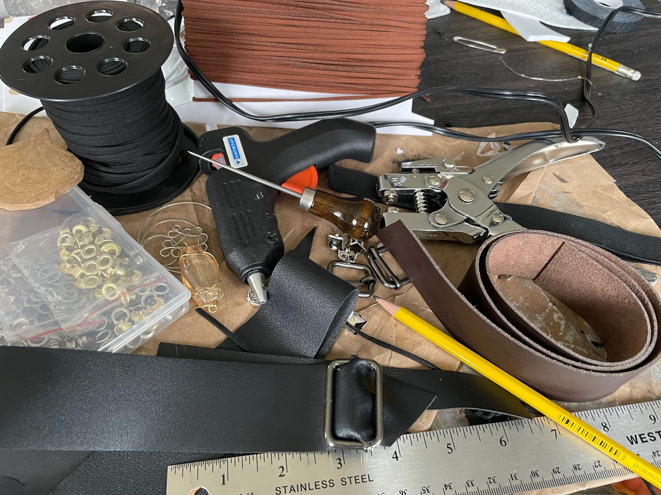 A cluttered workspace with various tools and supplies, including a spool of black gaskets, a glue gun, a staple remover, a yellow pencil, a measuring ruler, and some leather straps and cuts on a workbench.