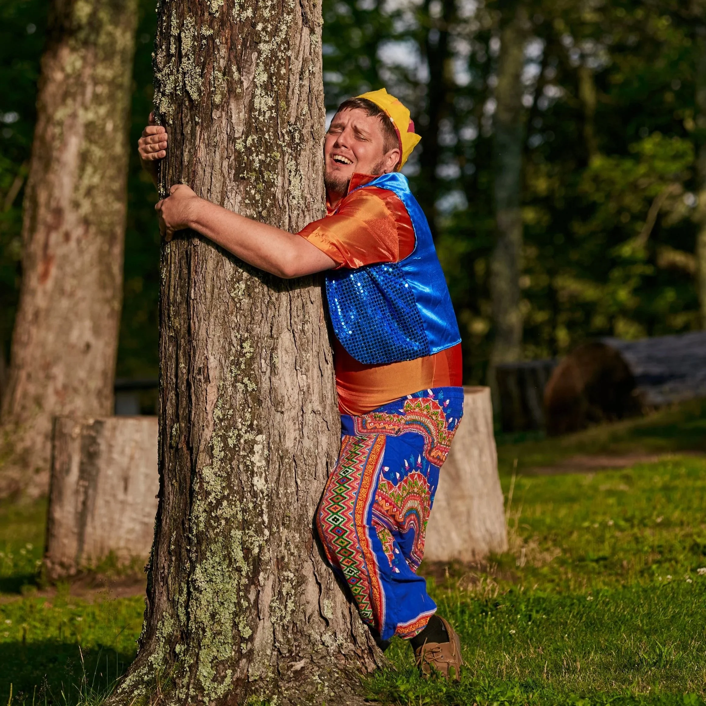 A man dressed as a clown is hugging a tree in a forest, with only his face, arms, and part of his costume visible. The background consists of green trees and grass.