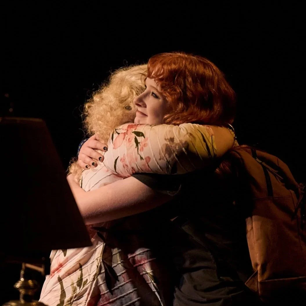 Two women hugging warmly, one with curly blonde hair and the other with straight red hair, in a dimly lit setting.