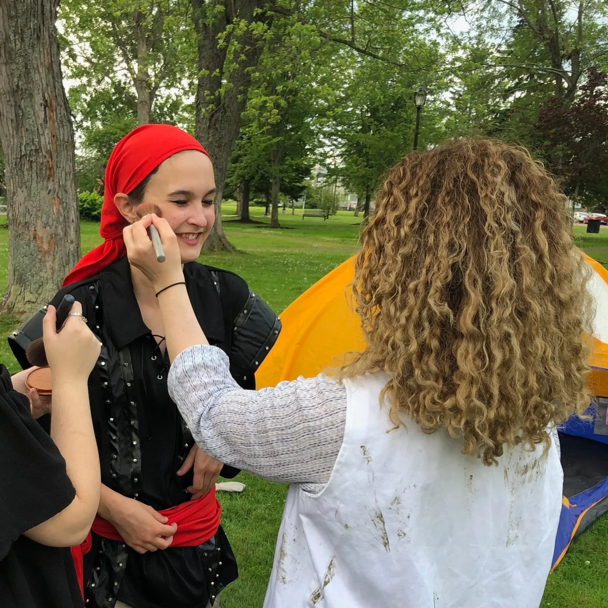Two women interacting outdoors in a park with trees and a grassy area, one with curly blonde hair and the other wearing a red headscarf and black jacket, smiling and holding makeup brushes.