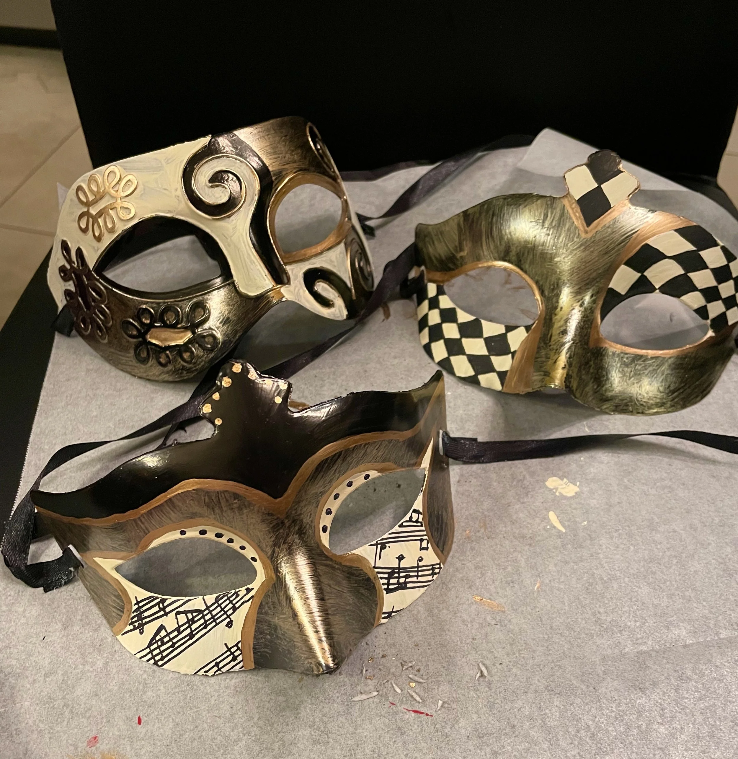 A collection of decorative masks with musical notes and pattern designs, resting on a table with a black cloth background.