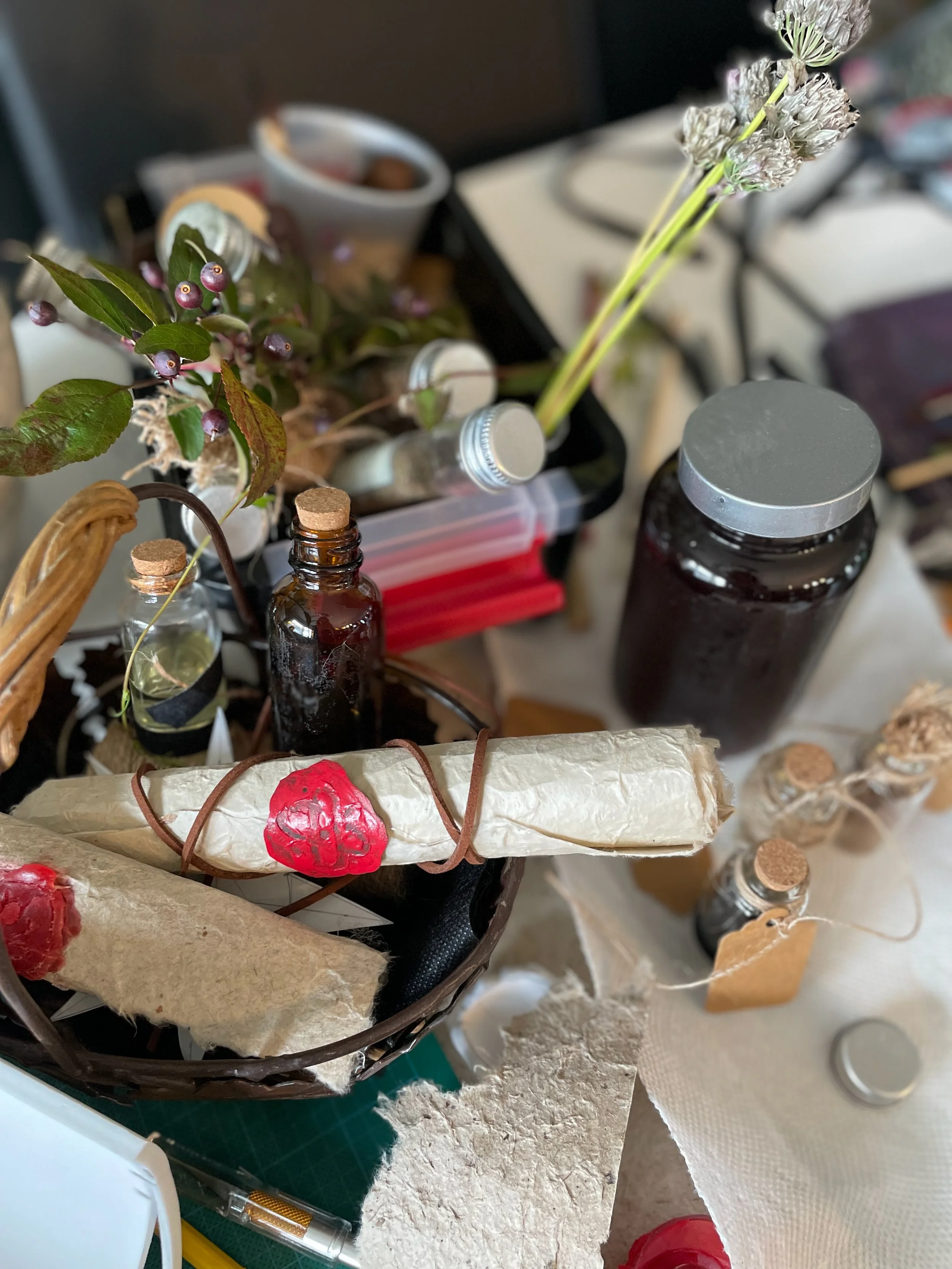 Various craft supplies and dried flowers arranged on a cluttered surface, including small bottles, paper-wrapped objects with red seals, and a tray with dried flowers and berries.