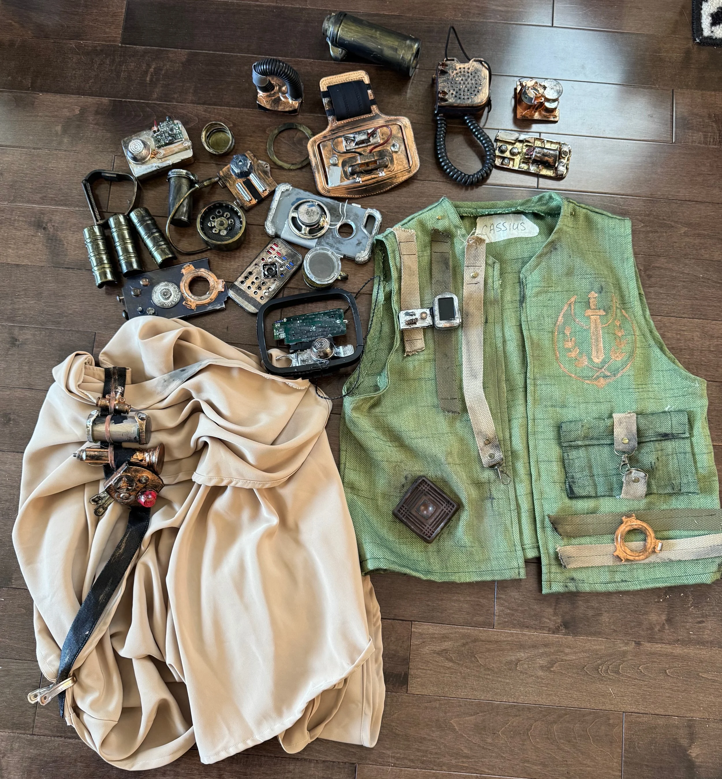 Various metallic and electronic parts scattered on a wooden floor, including wires, boxes, and mechanical components, along with a green cloth bag labeled 'CASSIUS' and a beige fabric item.