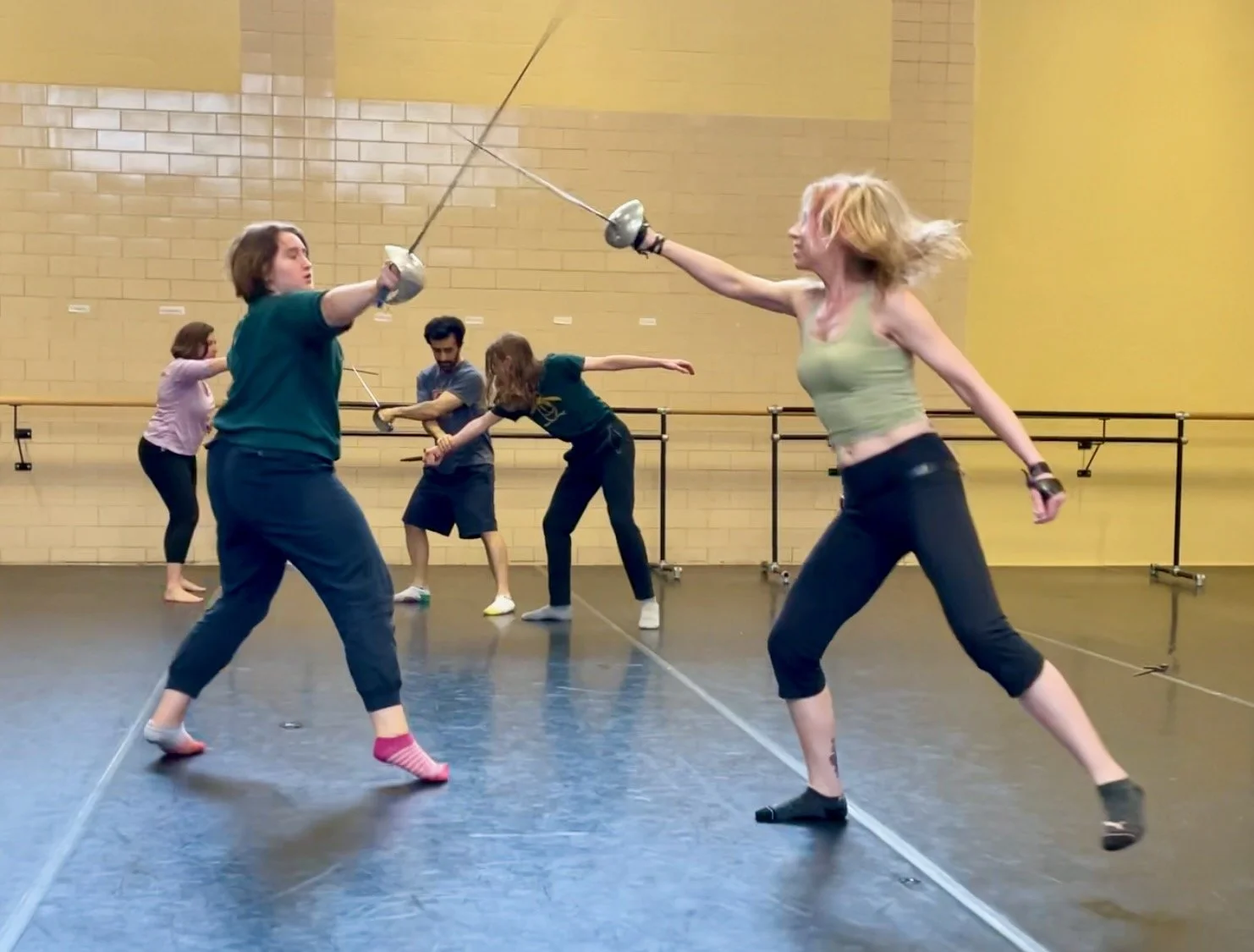 Two women practicing fencing in a dance studio with a yellow brick wall, others practicing in the background.