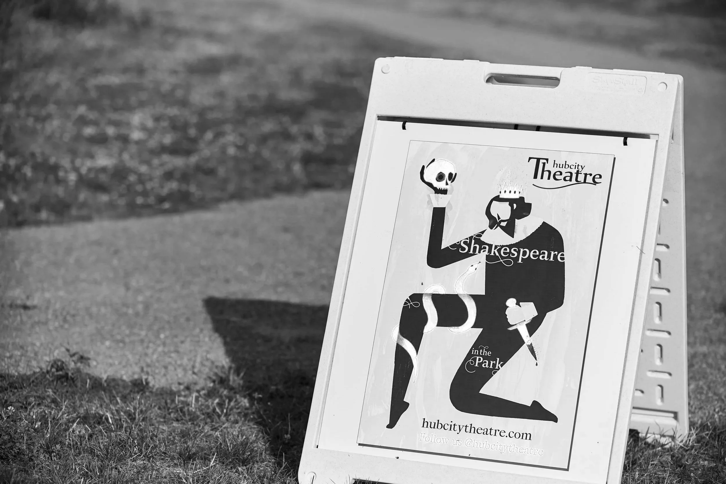 A black and white photograph of an A-frame sidewalk sign promoting the hubcity Theatre, featuring stylized illustrations of a skull and a theatrical mask, set outdoors on a grassy area with a blurred background.