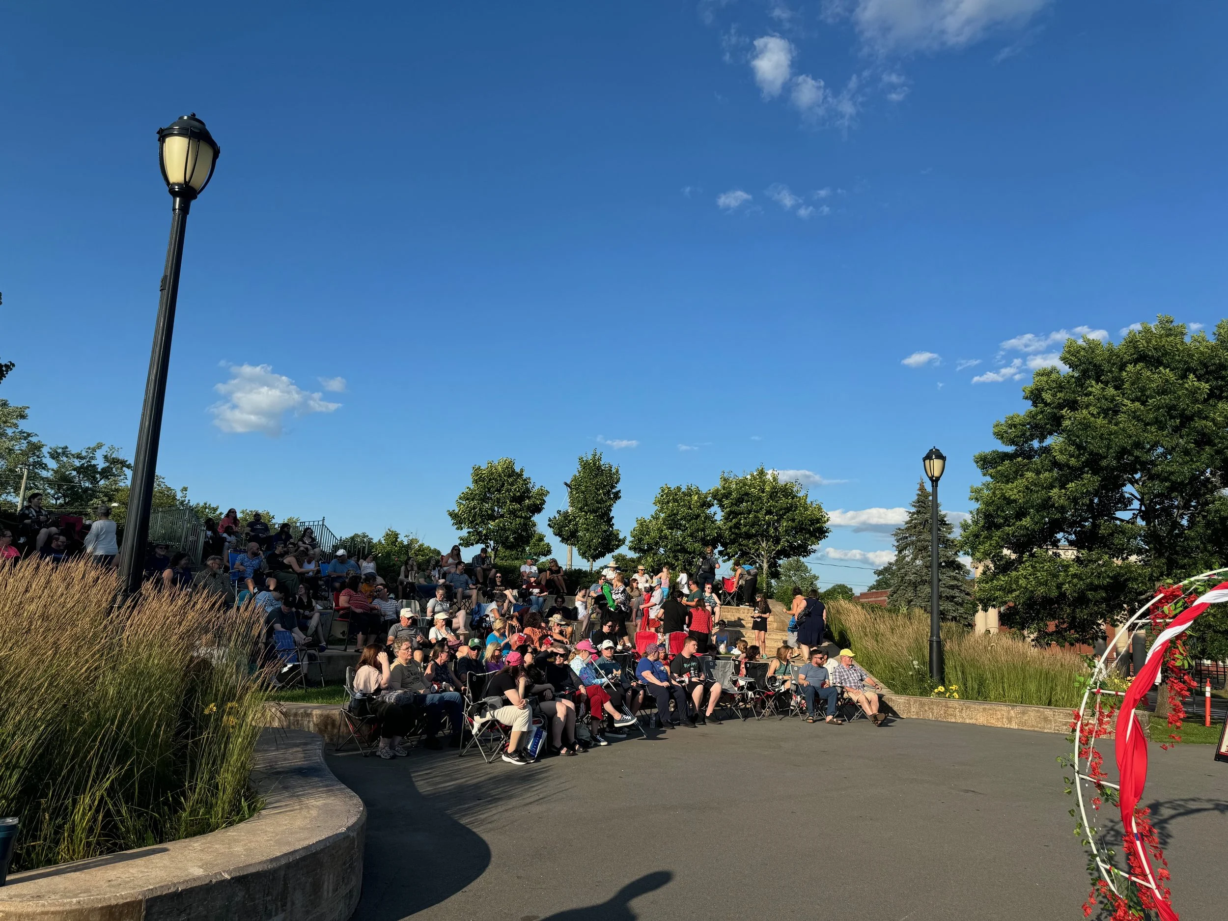 A large group of people seated outdoors on a sunny day, attending an event or gathering, with trees and a blue sky with scattered clouds in the background.