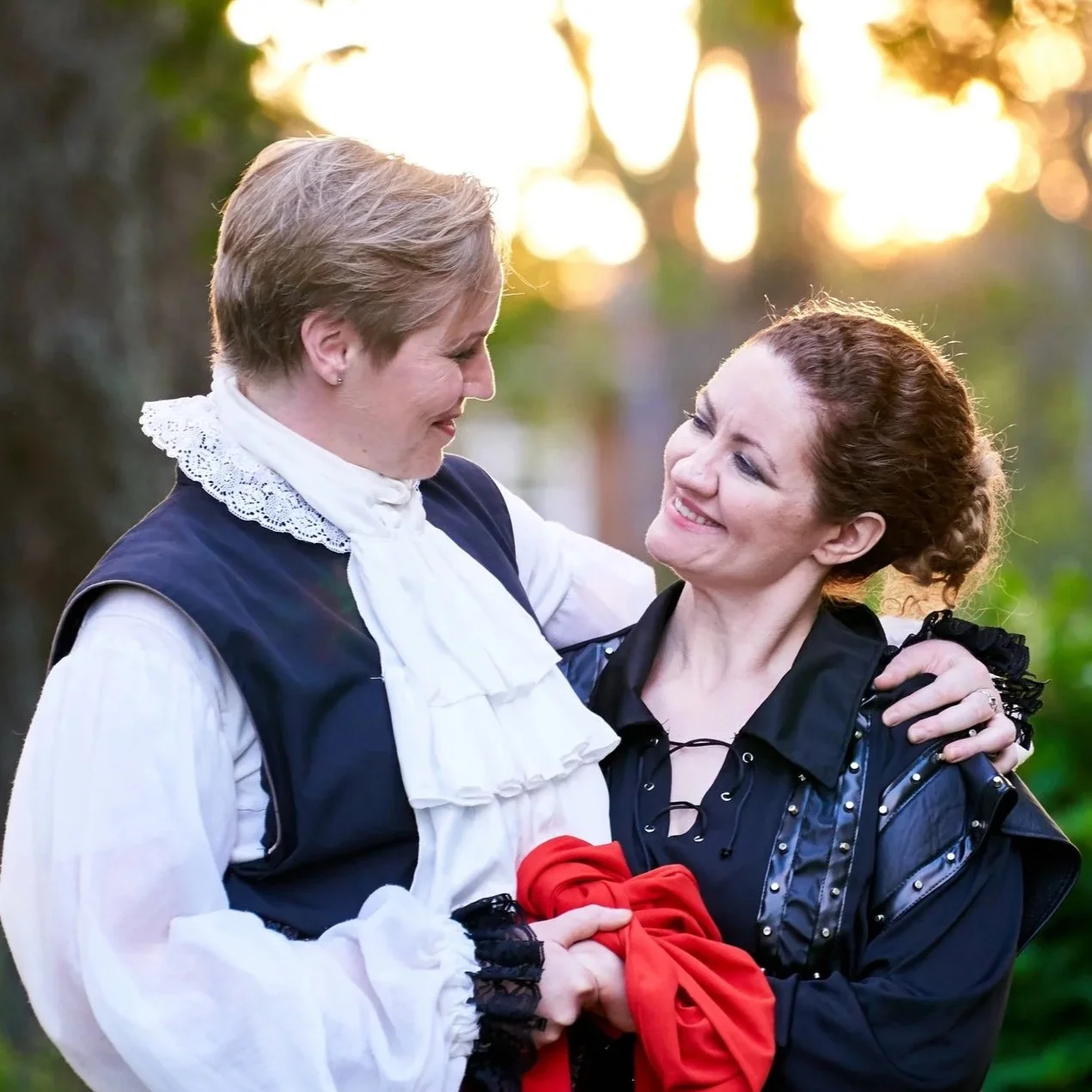 Two women smiling and holding hands outdoors during sunset, with one wearing a historical costume and the other in a modern dark outfit.