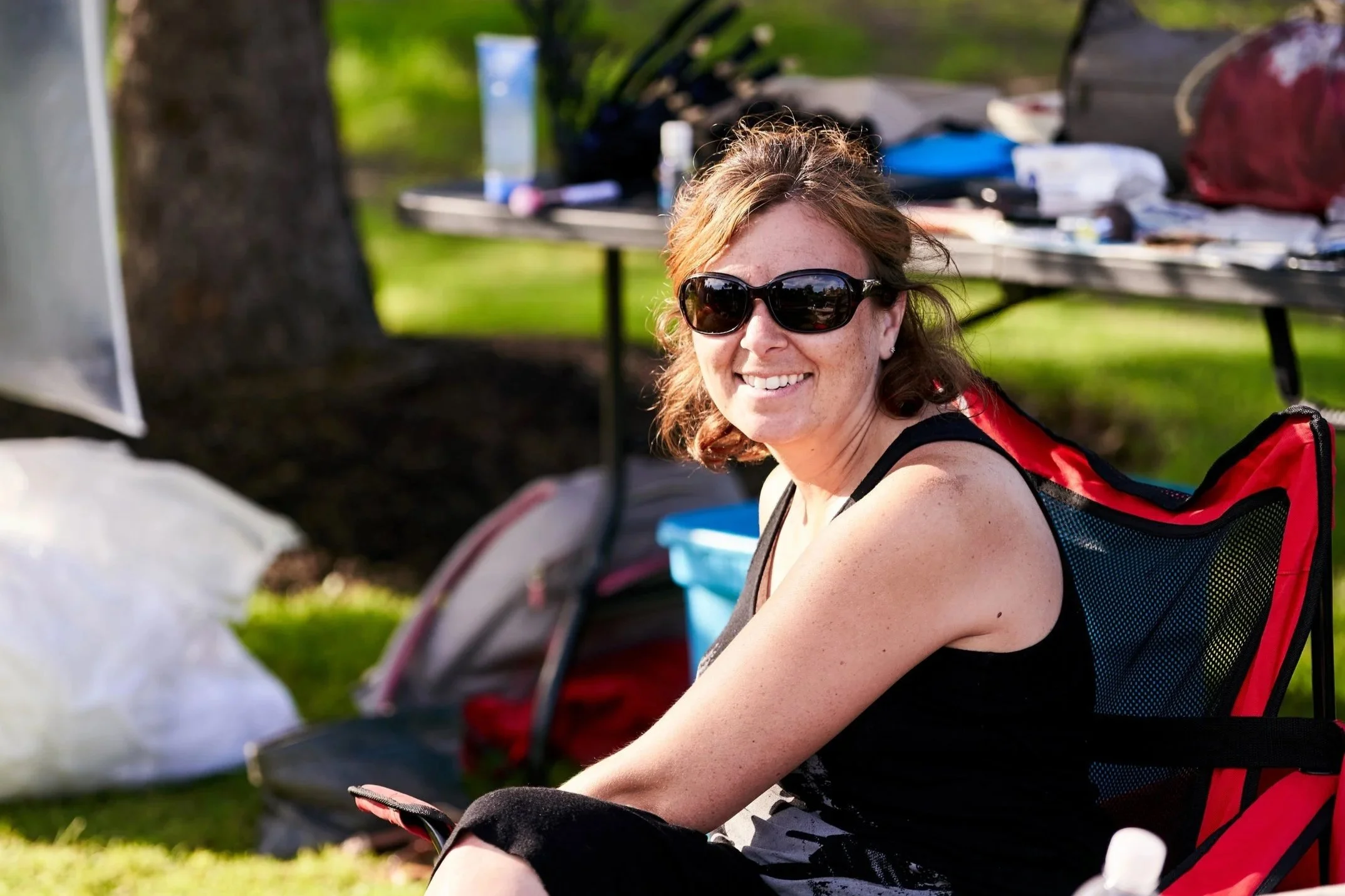 A woman with red hair wearing sunglasses, black sleeveless top, and a red backpack, sitting outdoors near a tree, with a picnic table and various items in the background.
