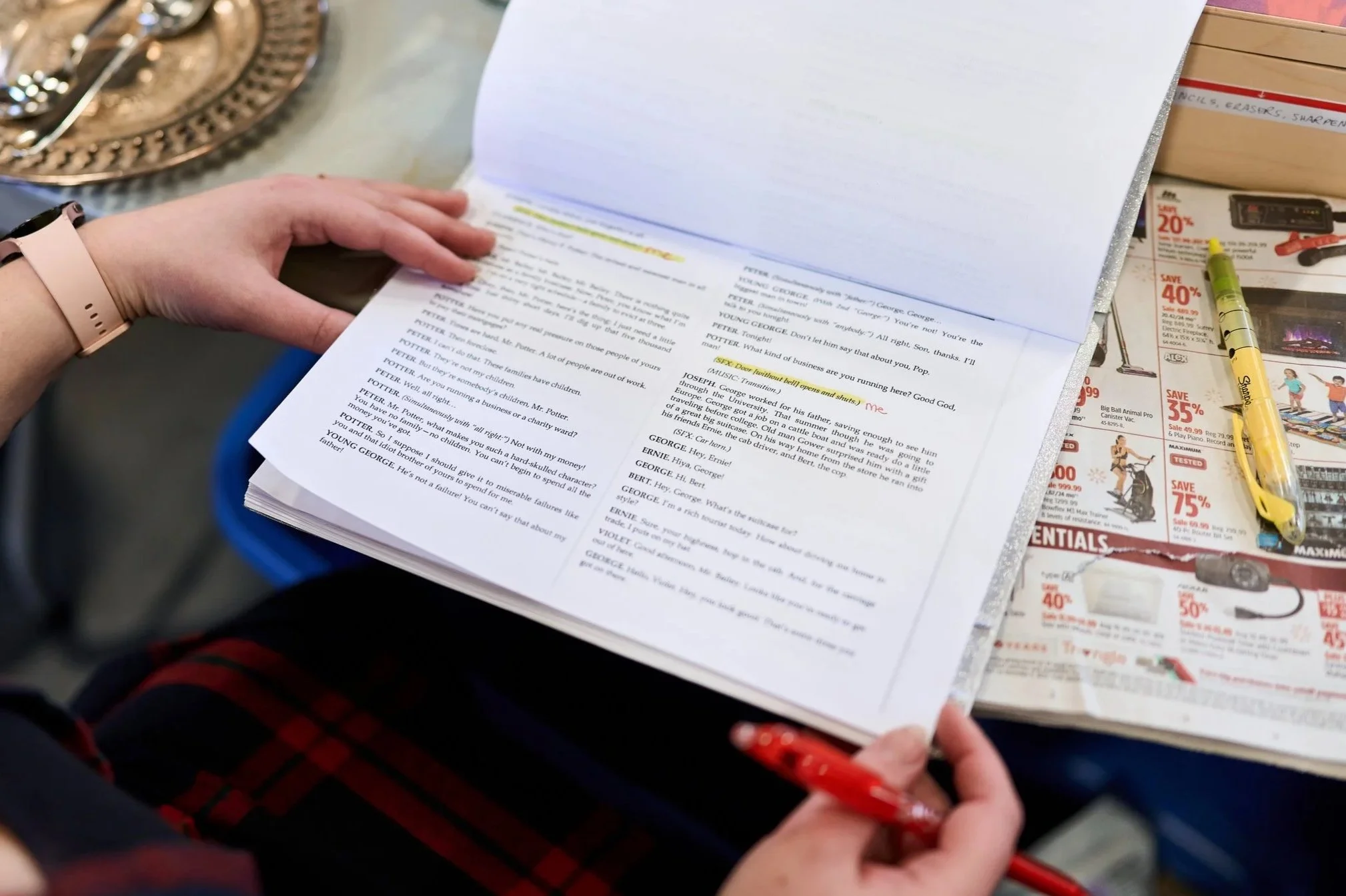 A person is holding and turning a script with highlighted lines, sitting at a table with a red pen in hand. The table has a metallic tray and various items, including a yellow pen, pages from a flyer or advertisement, and a box of colored pencils or similar art supplies.