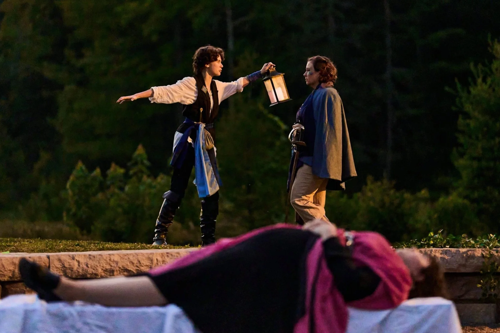 Two women dressed in historical pirate costumes stand outdoors at dusk, with trees in the background. One woman is holding a lantern and appears to be speaking to the other, who is listening.