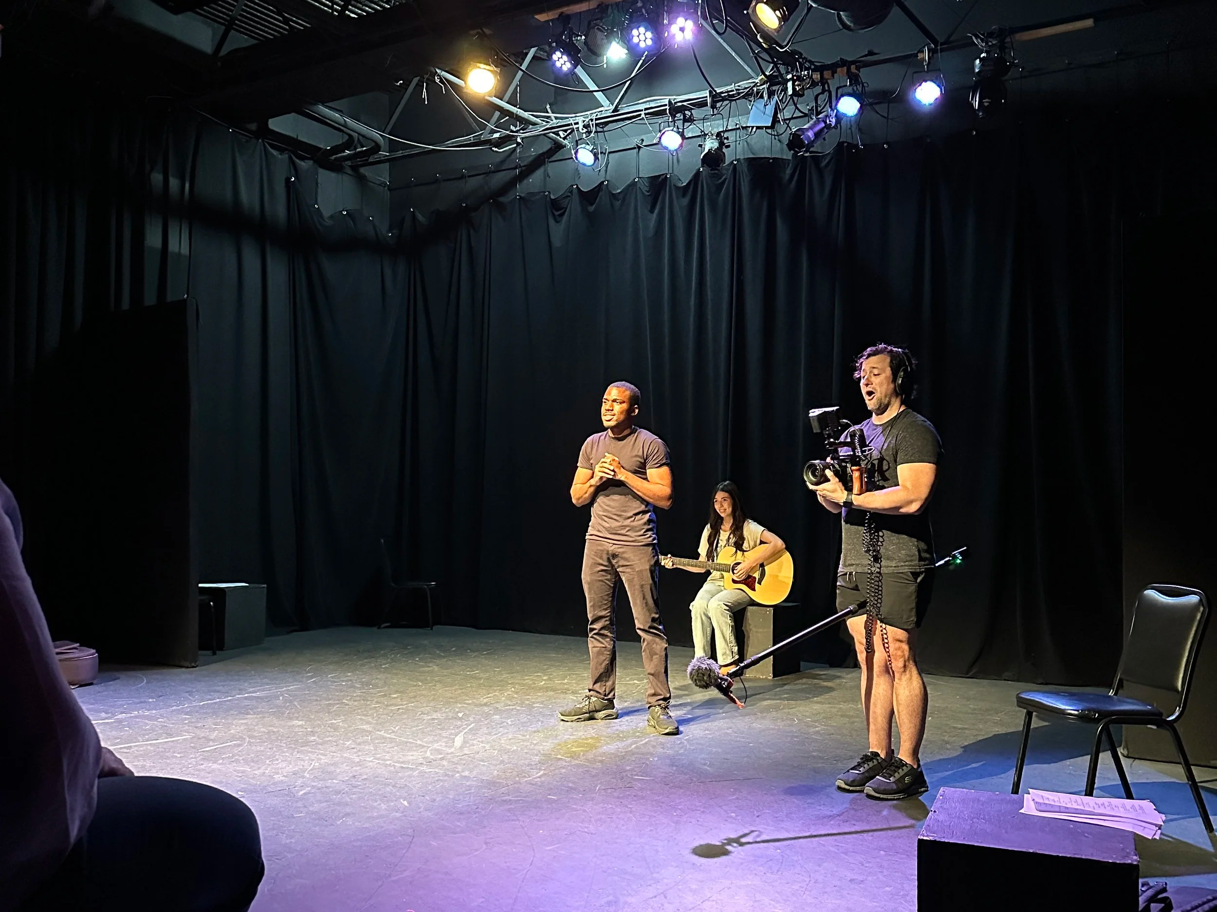 A stage with three performers: one man singing, a woman playing guitar, and another man filming with a camera. Black curtains and stage lights in the background.