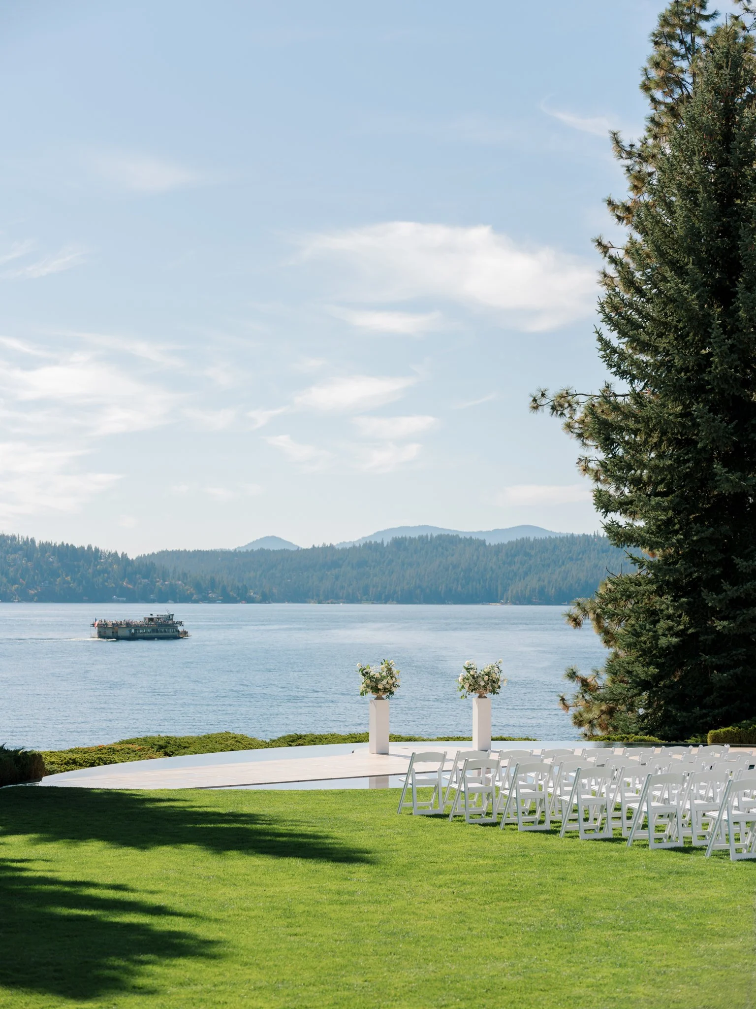 Outdoor wedding ceremony setup with white chairs and floral arrangements near a lake with a ferry and mountains in the background at the Hagadone Event Center in Coeur d'Alene, Idaho.